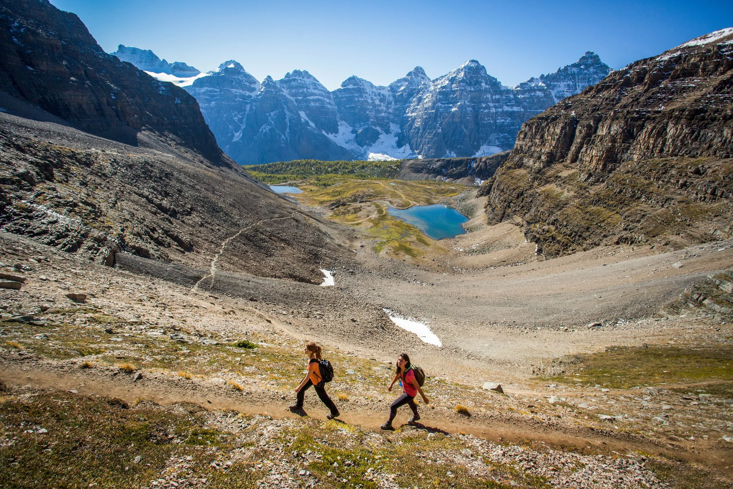 Two women in hiking gear walking along the rocky shoreline of a turquoise alpine lake with snow-capped mountain peaks in the background.
