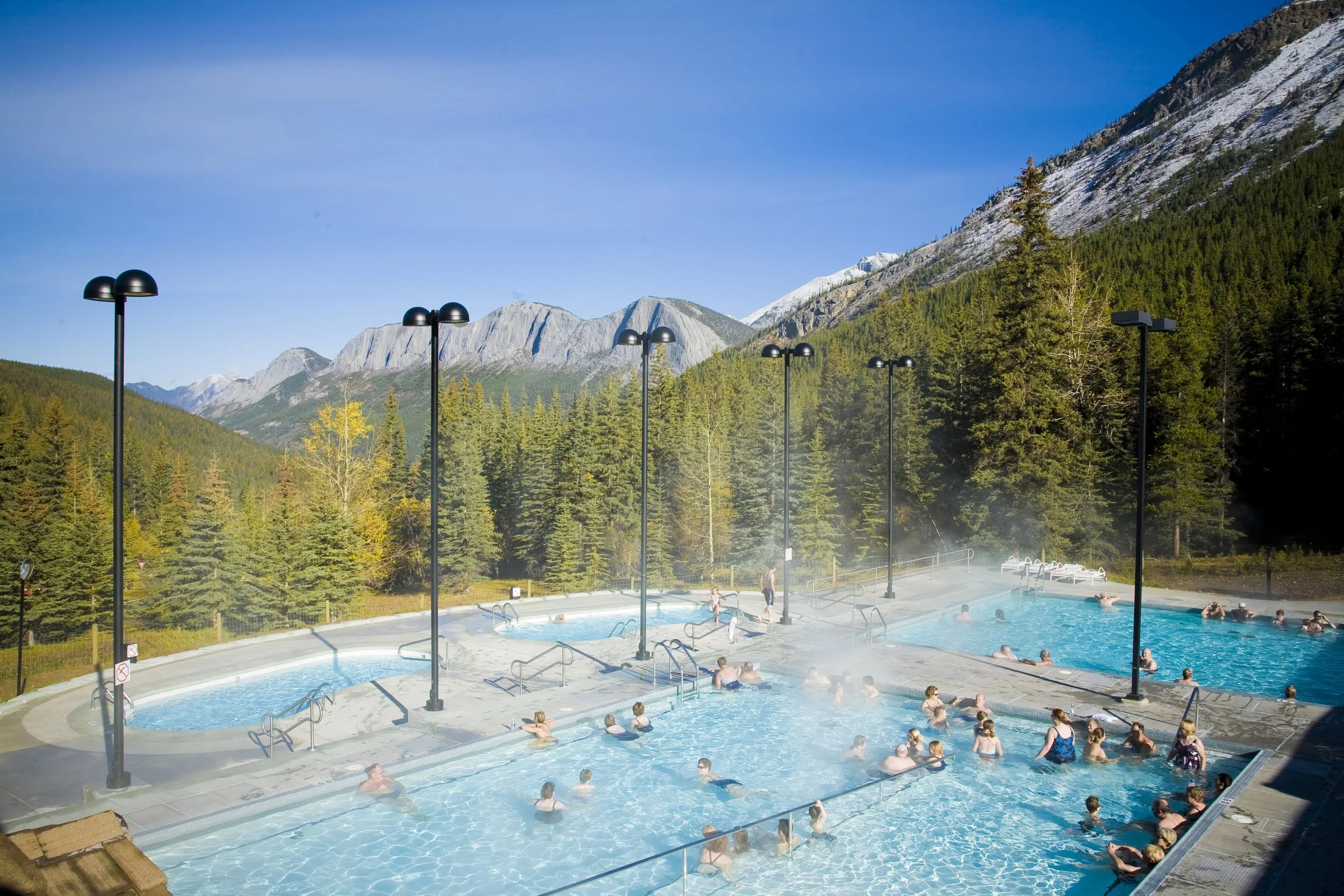 Visitors swimming in large outdoor thermal hot spring pools surrounded by lush pine trees and a dramatic mountain backdrop under a clear blue sky.