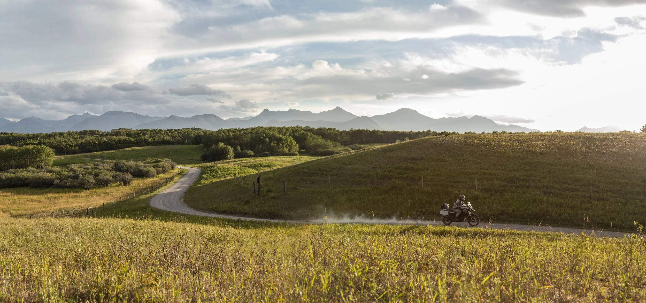 Wide-angle shot of a motorcyclist riding down a winding gravel road through rolling green foothills, kicking up dust against a backdrop of distant jagged mountain peaks under a dramatic, cloudy sky.