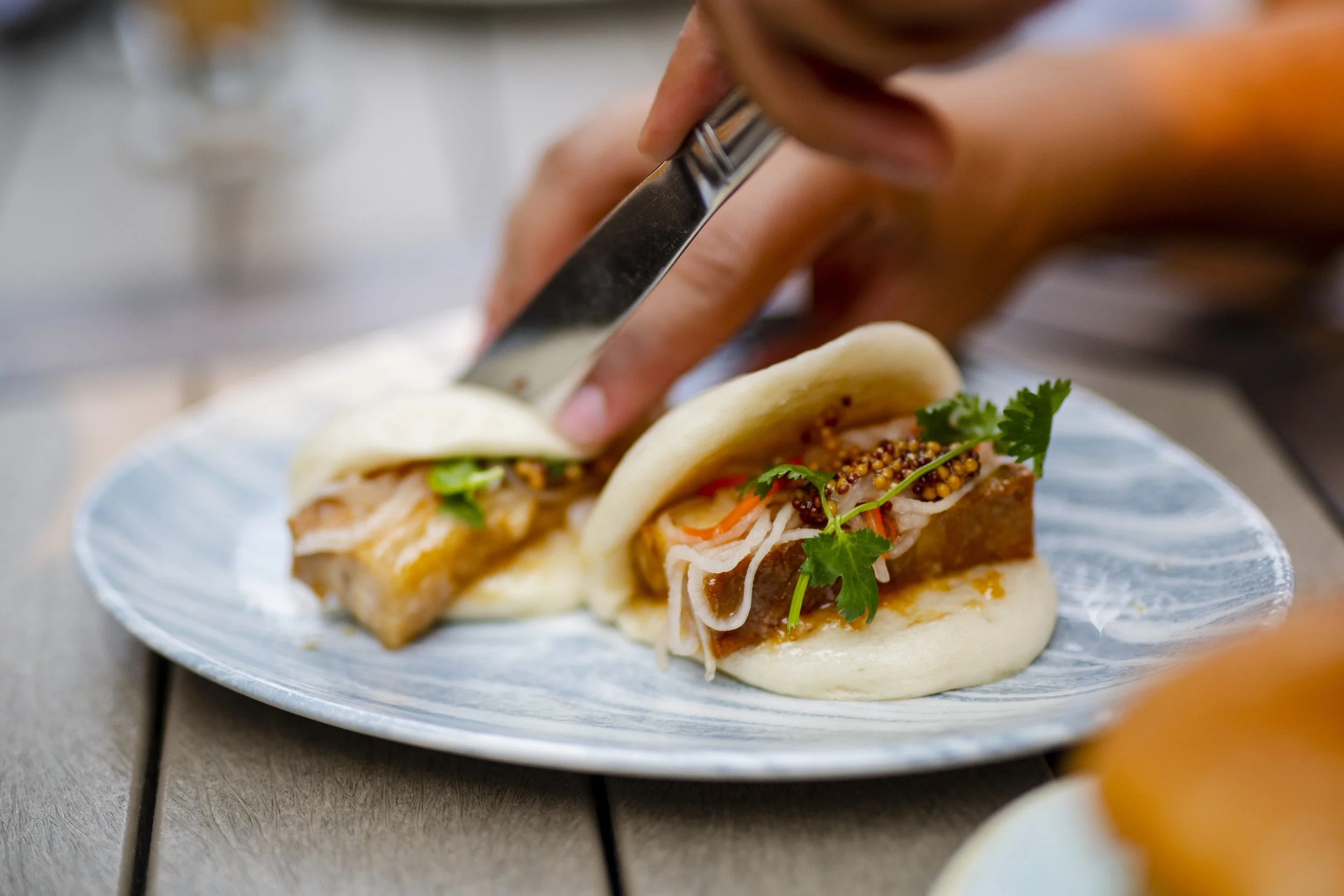 Close-up of a person using a knife to slice a gourmet pork belly bao bun garnished with mustard seeds and fresh cilantro on a blue marbled plate.