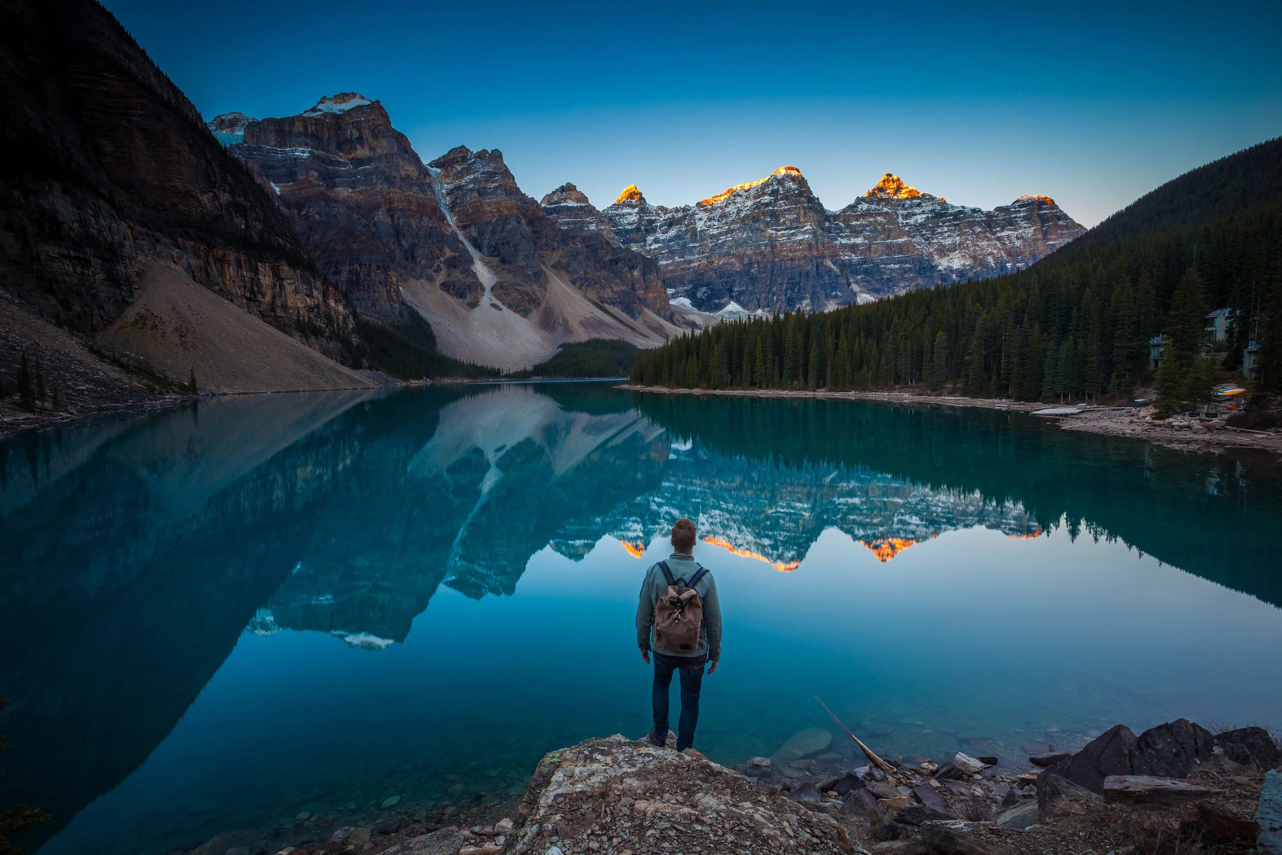 A hiker with backpack trekking up a steep rocky mountain trail overlooking a scenic valley with alpine lakes and rugged, snow-dusted peaks.