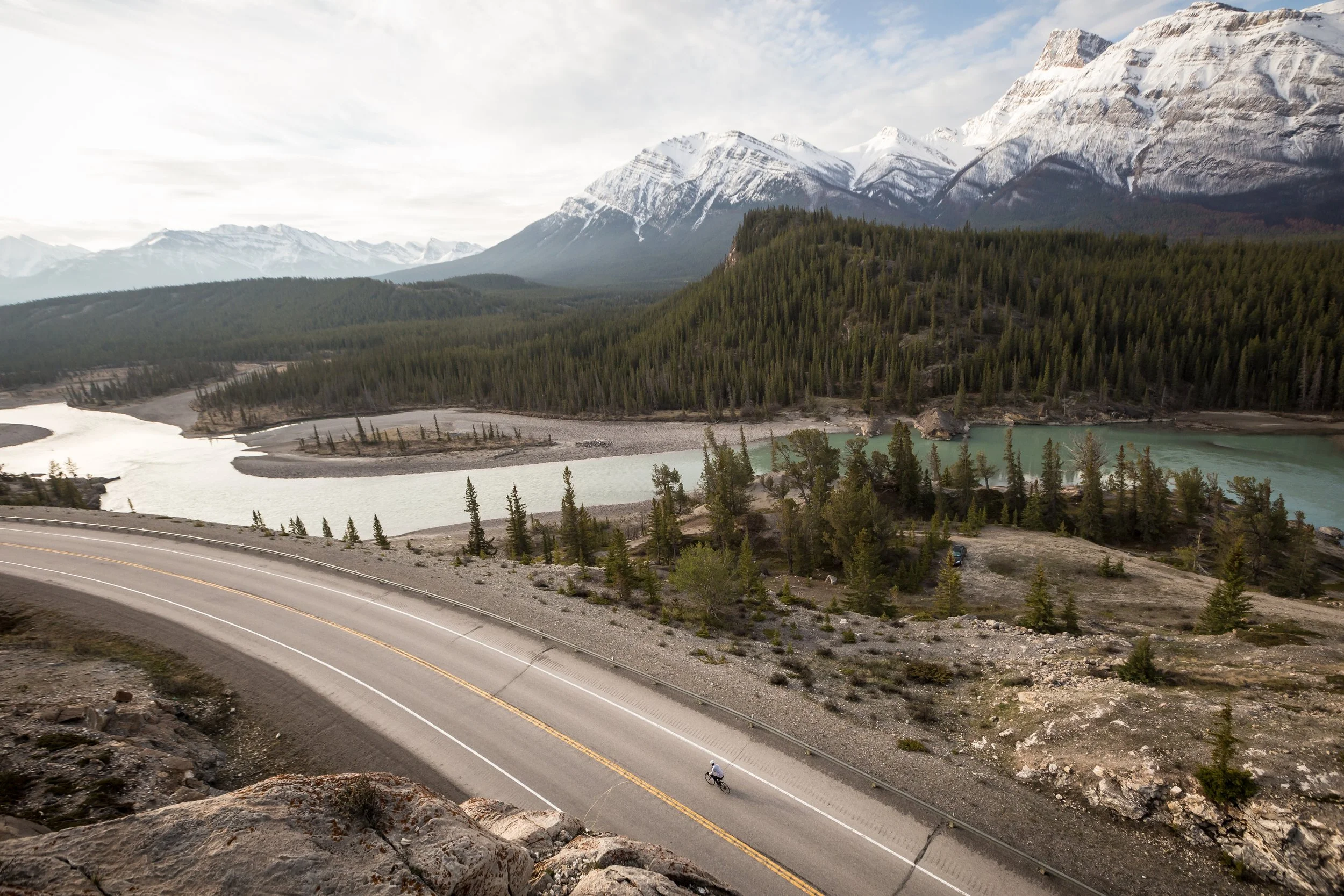 High-angle shot of a lone cyclist riding along a winding mountain highway in Banff parallel to a turquoise glacial river and dense evergreen forests in a vast mountain valley.