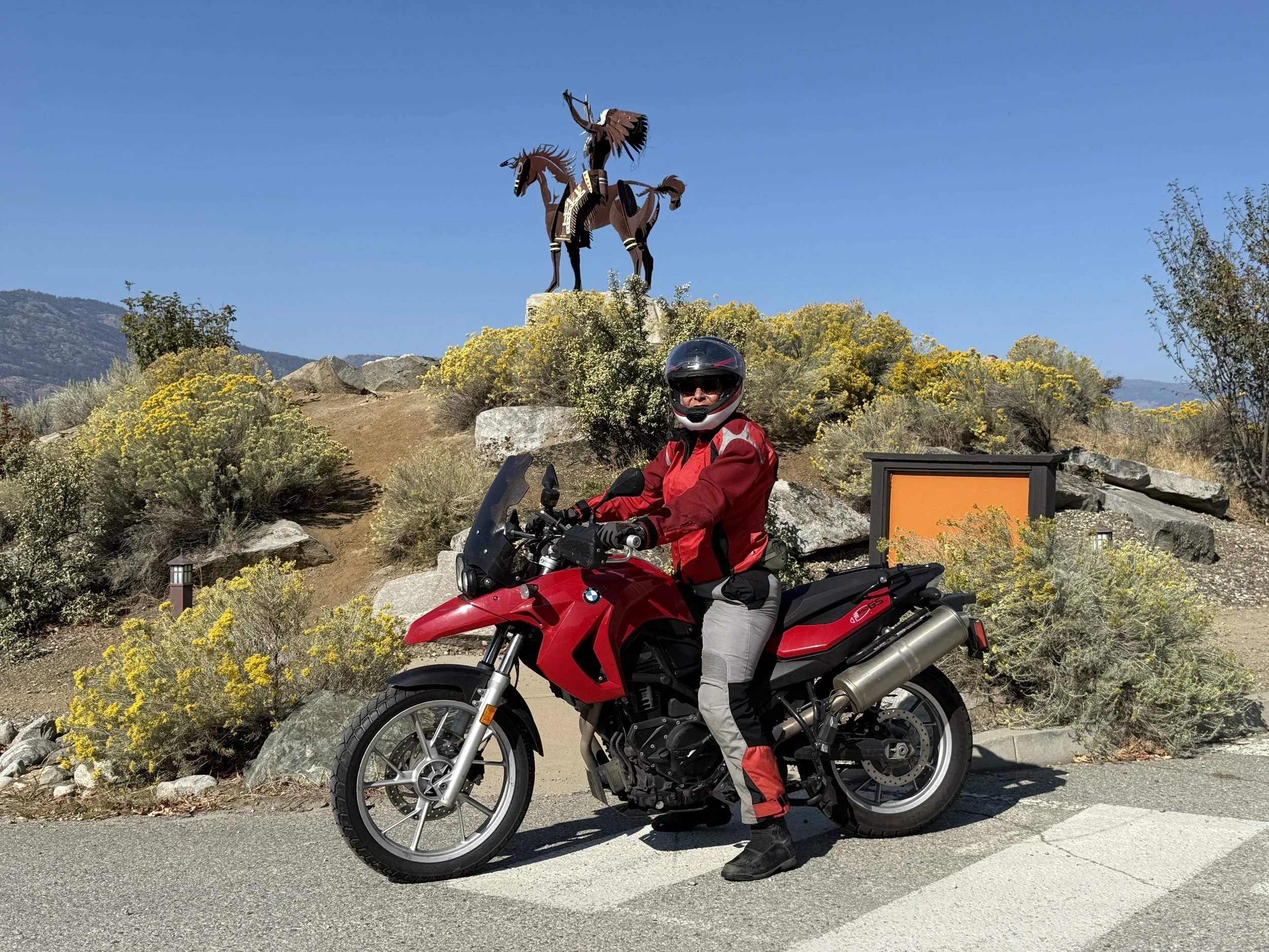 A woman in a red riding jacket and helmet sits on a red BMW GS adventure motorcycle parked on the side of a road in front of an Indigenous warrior on horseback statue.