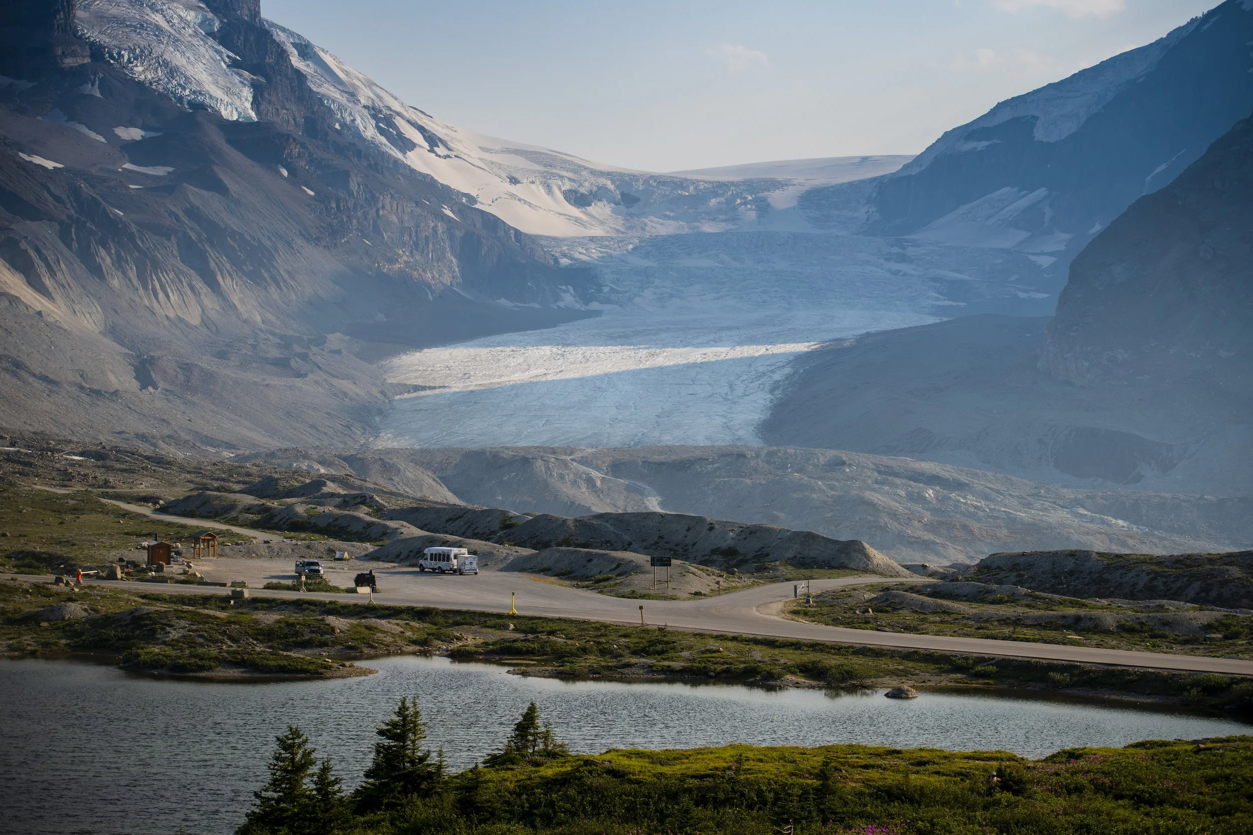 Wide landscape view of a massive alpine glacier nestled between rocky mountain peaks, with a small parking lot, road, and a calm lake in the foreground under soft daylight.