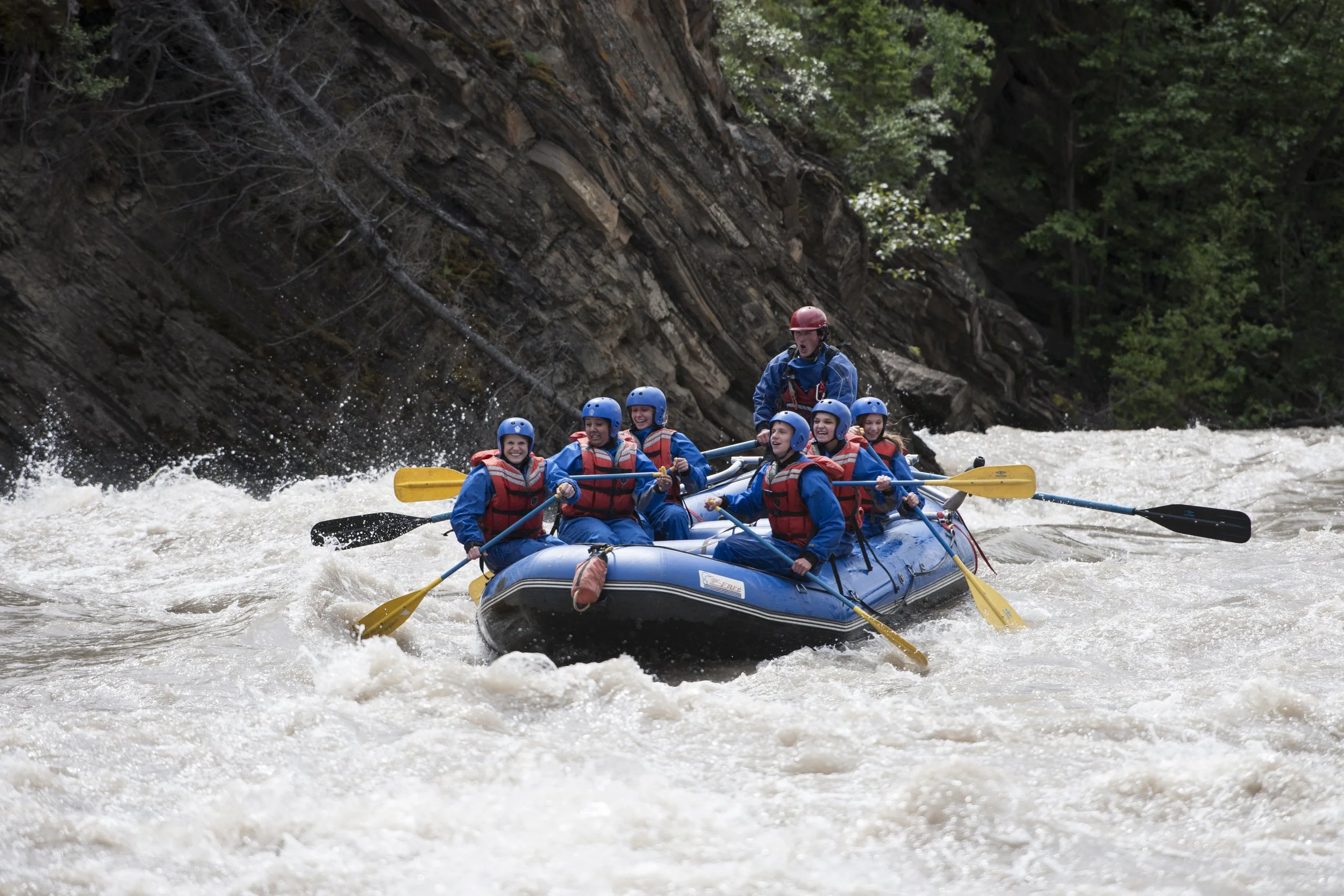 A group of people in blue helmets and red life jackets white water rafting down a rushing river alongside a steep rock wall.