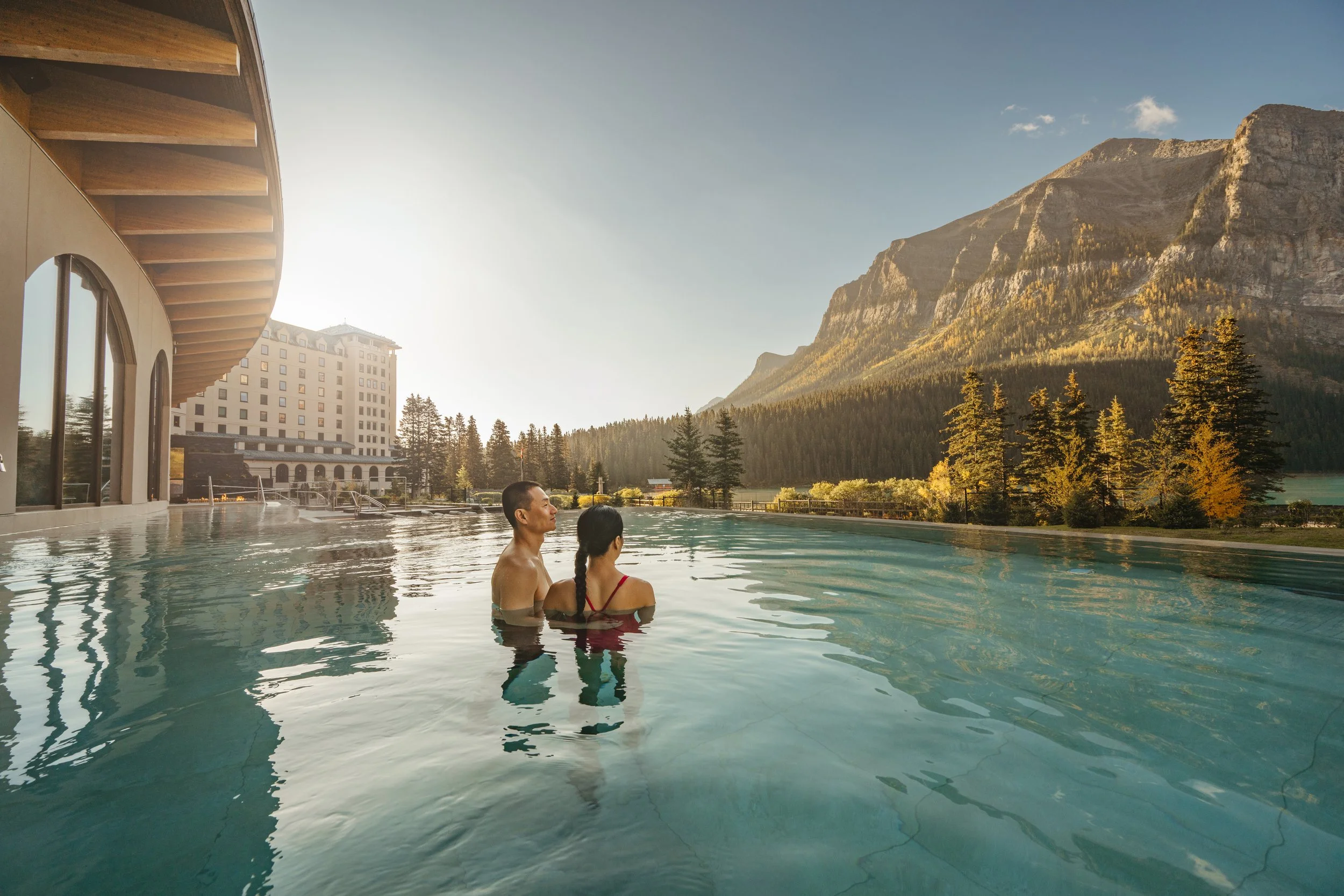 A couple relaxing in an outdoor infinity pool overlooking a serene mountain lake and steep rocky cliffs during a bright morning sunrise.