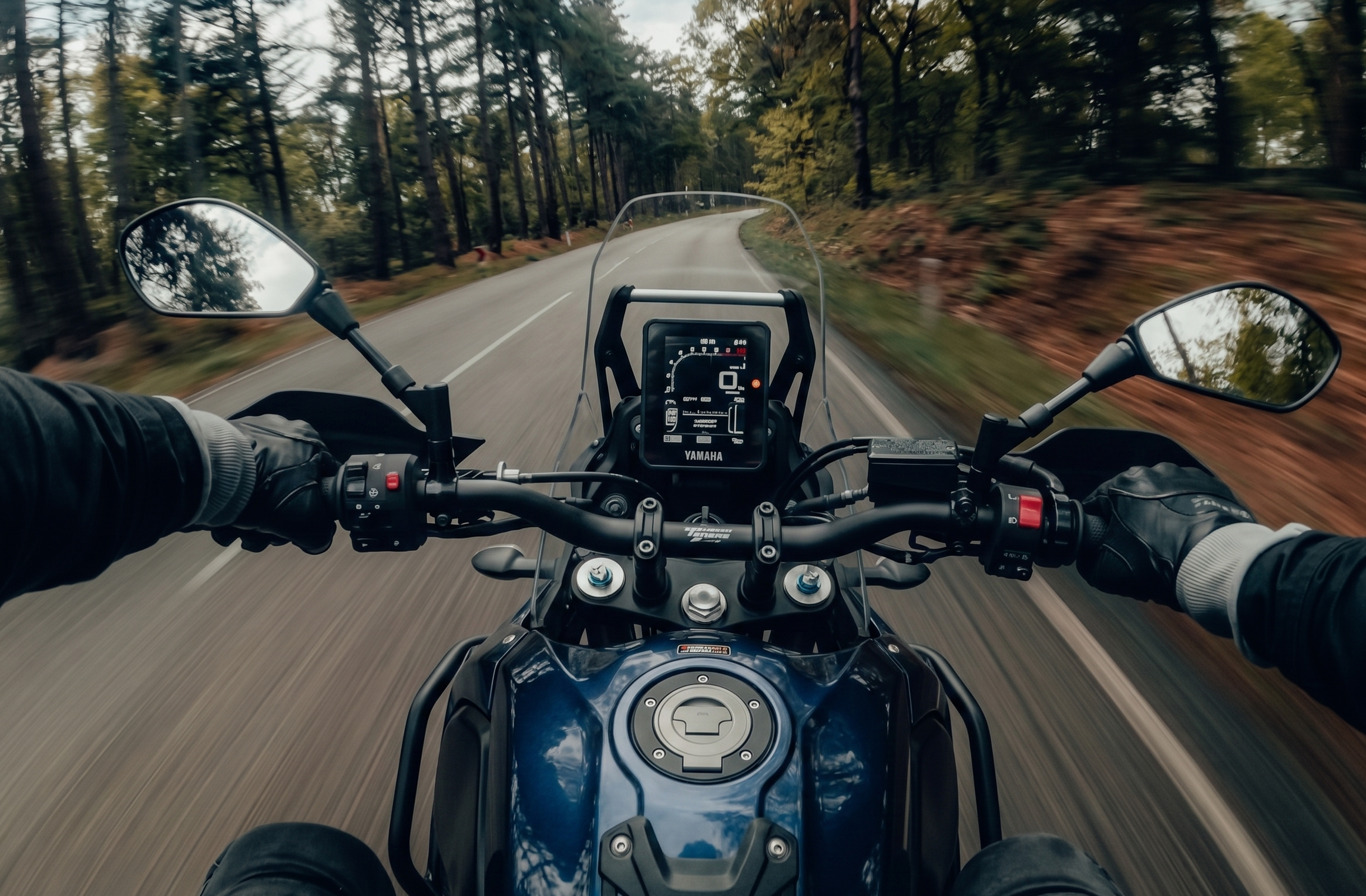 Point of view perspective of a rider on a blue Yamaha Tenere adventure motorcycle with a tall windscreen and digital dash, riding on a paved road through a forest.