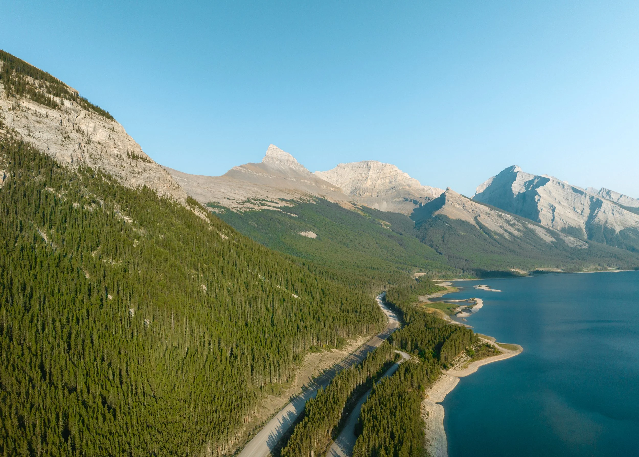 Aerial panoramic view of a winding mountain highway stretching between a dense evergreen forest and a deep blue alpine lake, set against a backdrop of rugged, sunlit limestone peaks under a clear sky.