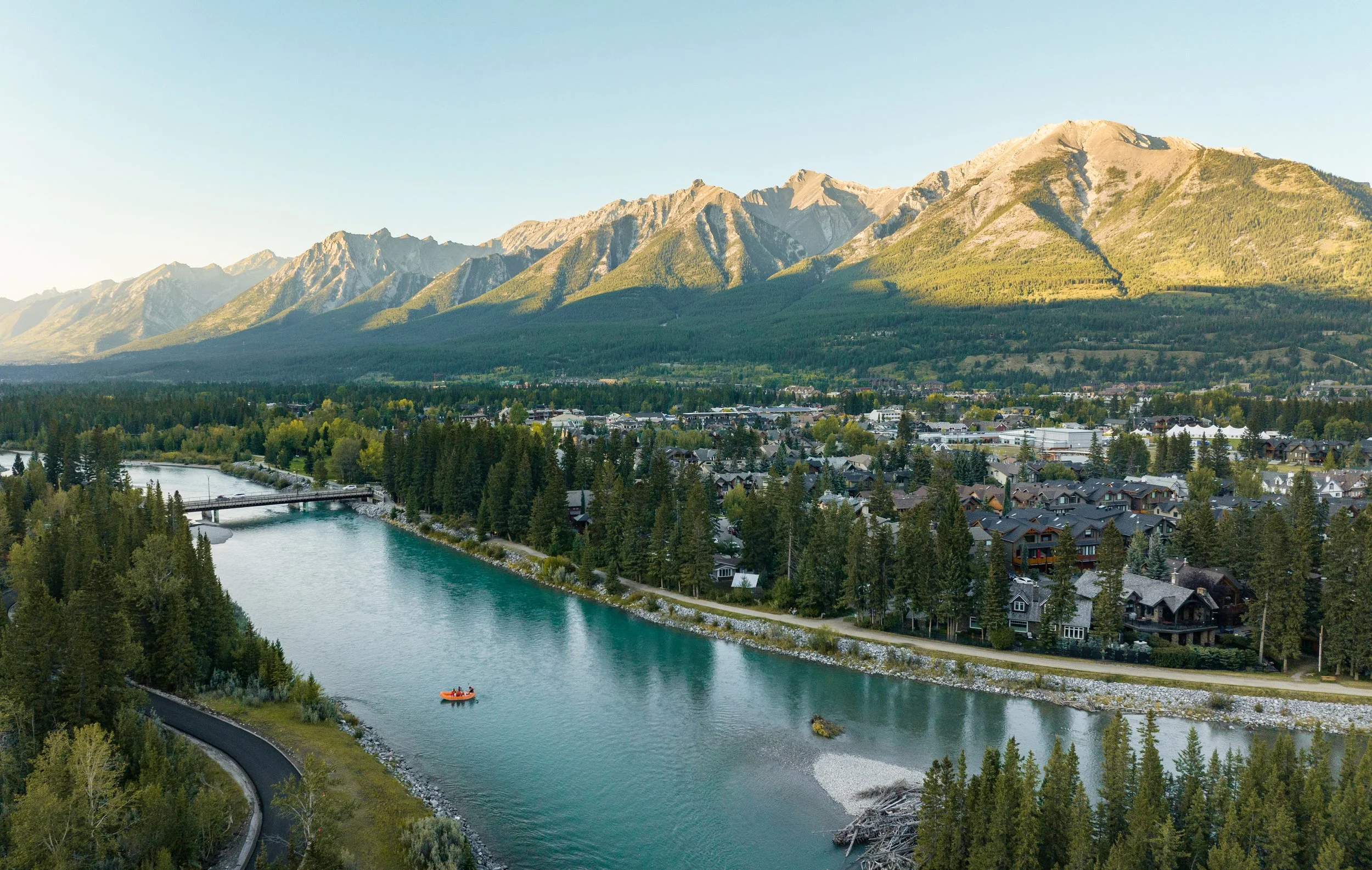 Wide-angle aerial view of the town of Canmore, Alberta, featuring the turquoise Bow River with a small orange raft, nestled against a backdrop of the majestic, sun-drenched peaks of the Canadian Rockies under a clear sky.