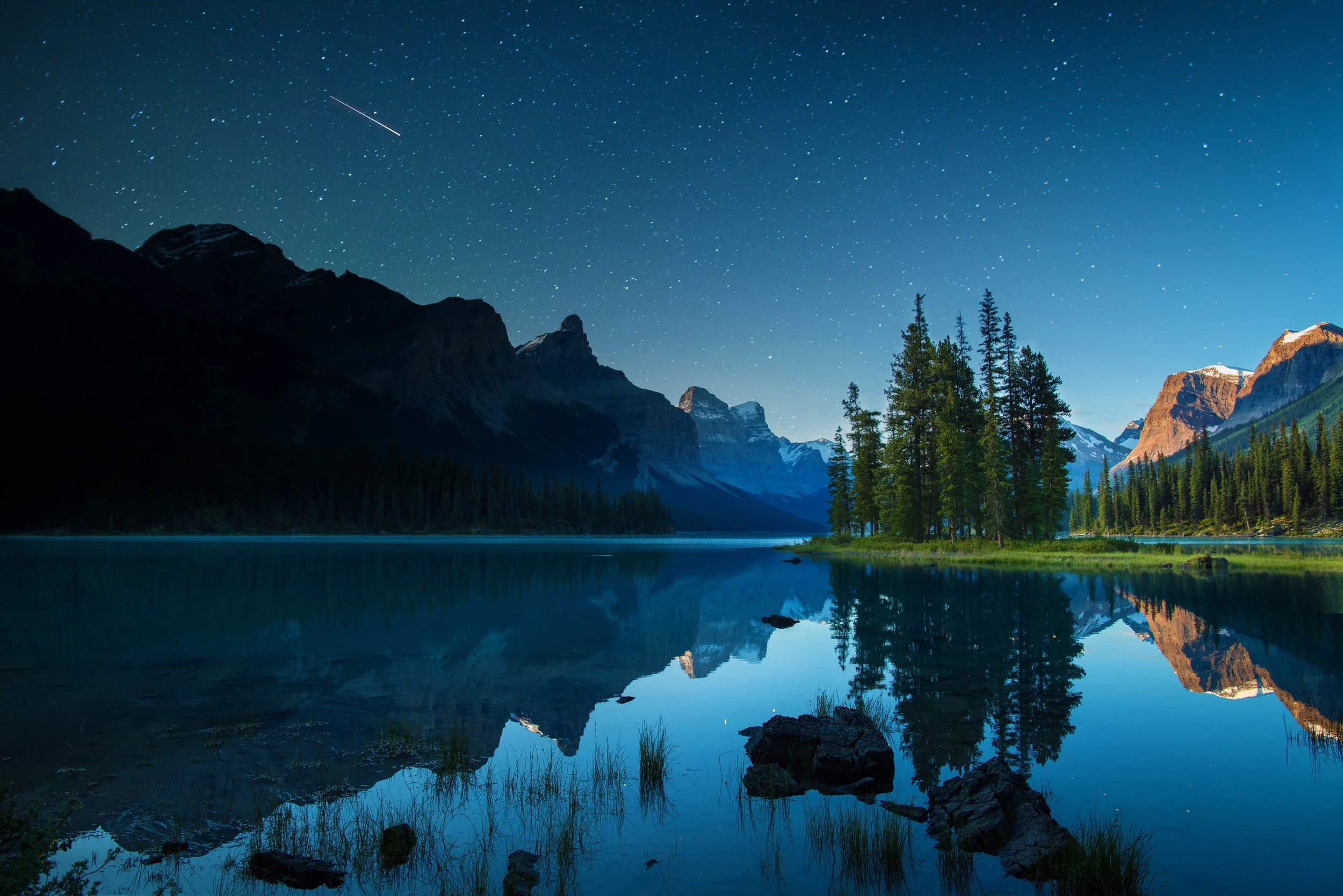 A starry night sky with a shooting star over Spirit Island on Maligne Lake, with perfectly calm water reflecting the surrounding pine trees and dark mountain silhouettes.