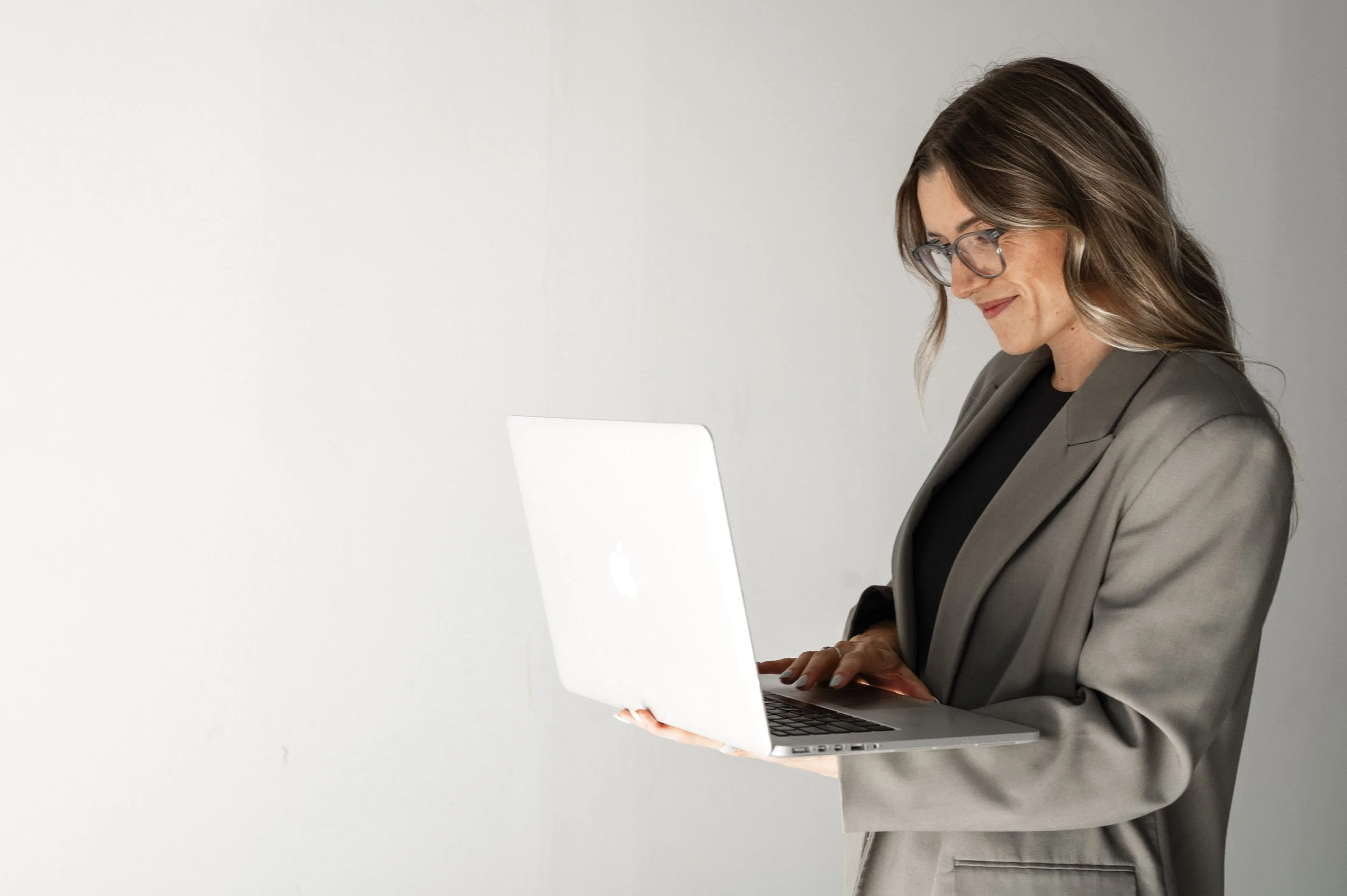 A woman wearing glasses and a gray blazer looks at a laptop screen with a slight smile against a plain white background.