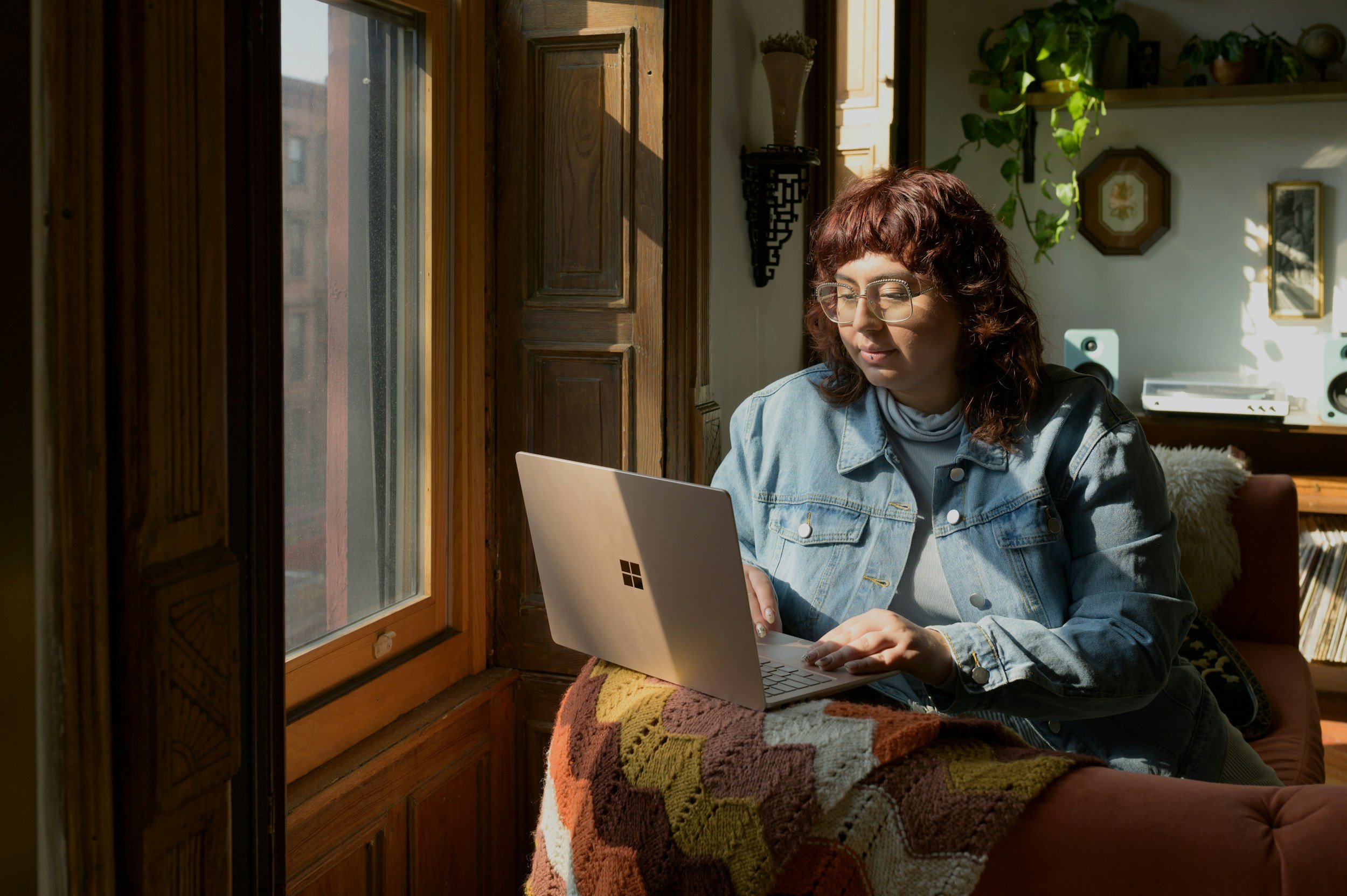A woman with glasses and curly hair sitting on a couch using a Microsoft Surface laptop, in a room with wooden window frames and sunlight.