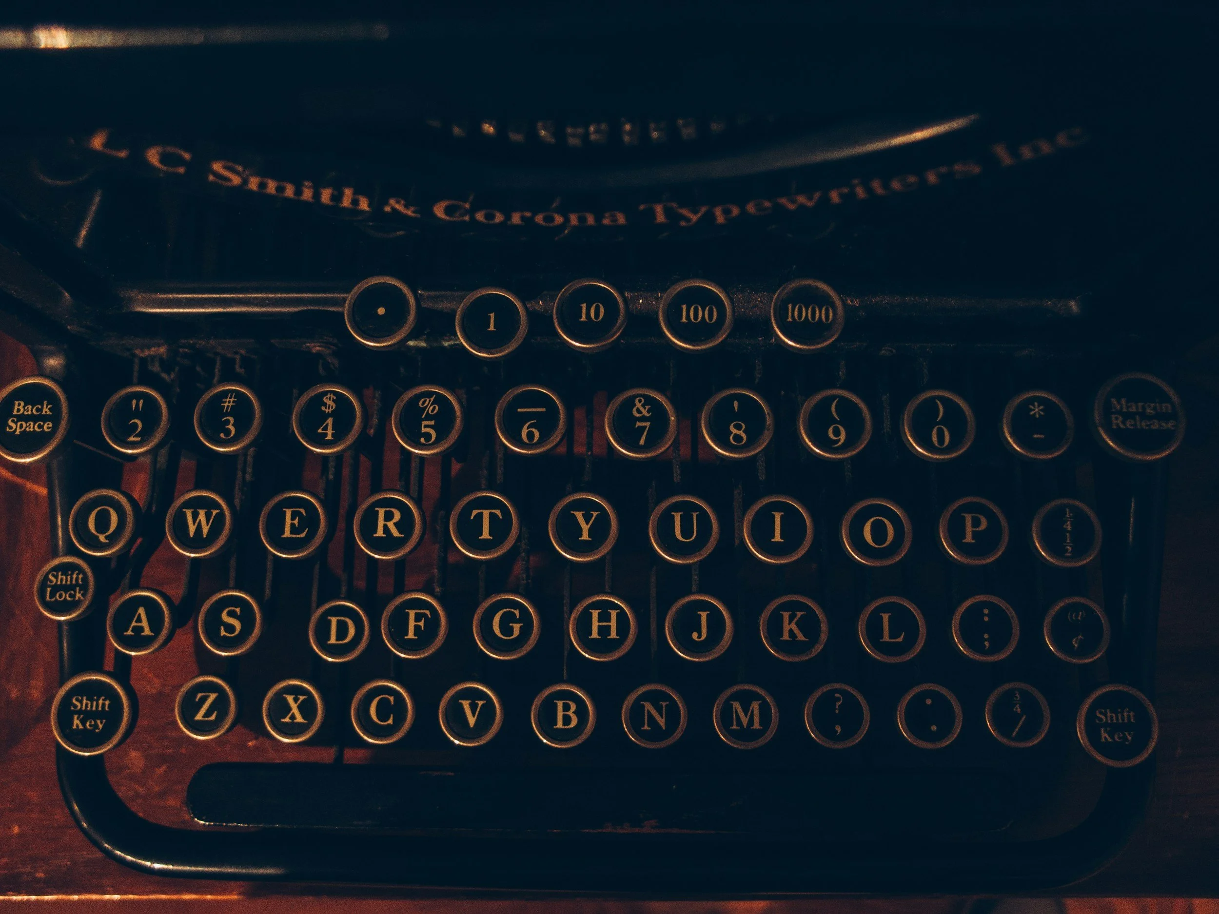 Close-up view of a vintage typewriter keyboard with round black keys and gold lettering. The visible keys include numbers 1, 10, 100, and 1000, along with letters Q, W, E, R, T, Y, U, I, O, P, and some symbols. The background shows part of the typewriter's body and typing mechanism.