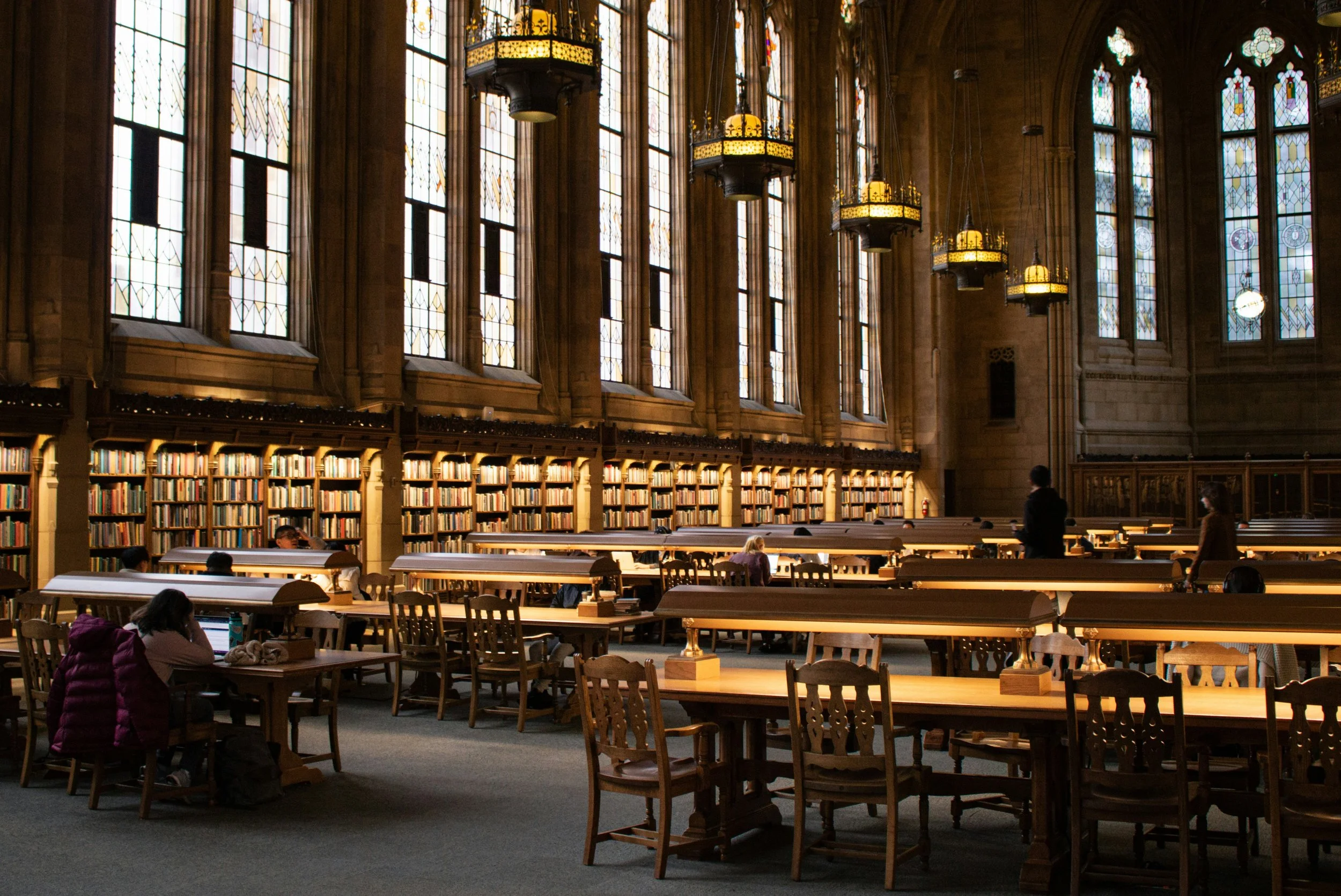 Interior of a historic library with large stained glass windows, wooden bookshelves filled with books, and wooden tables with chairs where people are reading or studying.