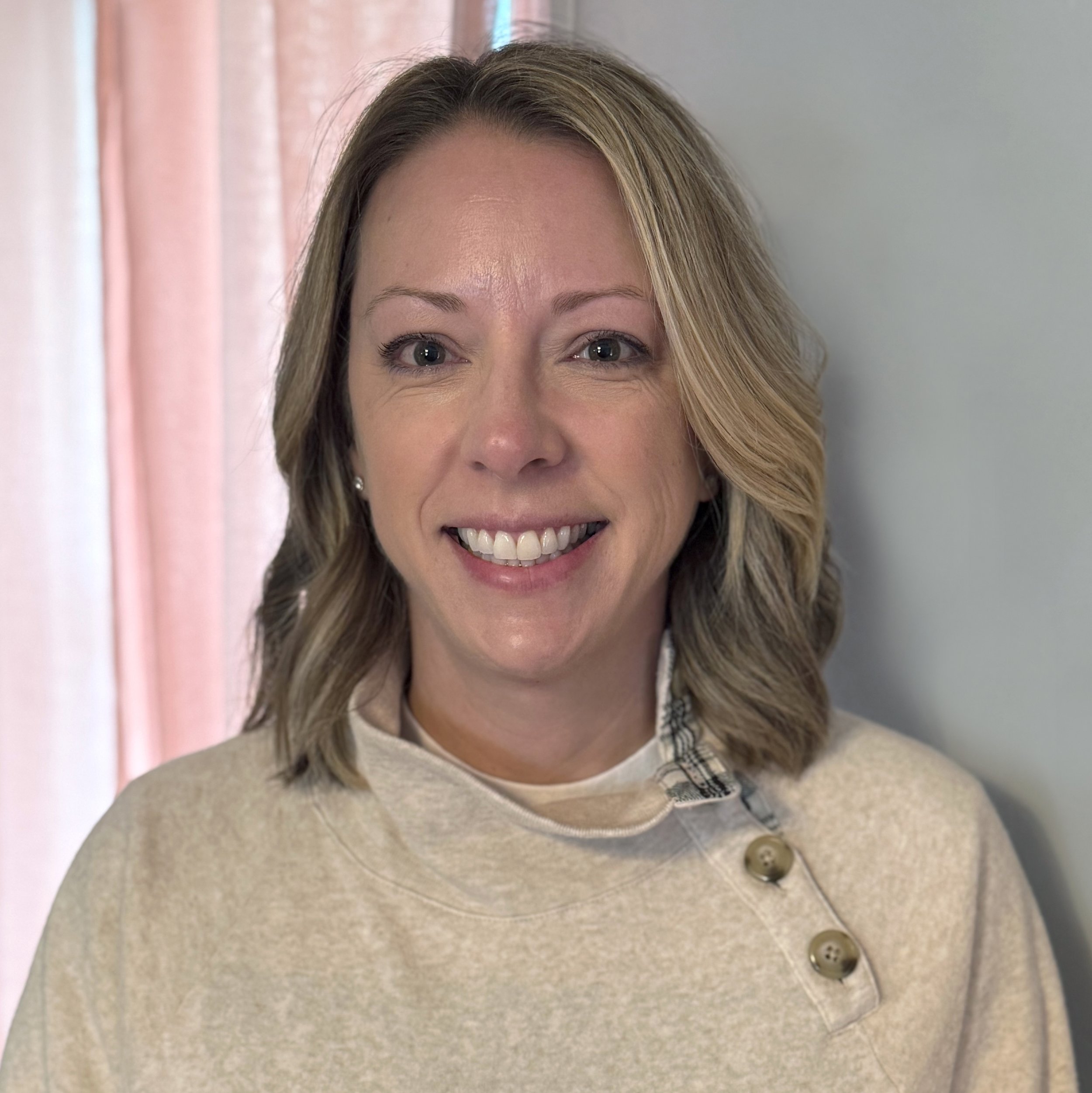 A woman with shoulder-length blonde hair, smiling, wearing a beige sweater with decorative buttons, standing against a light-colored wall near pink curtains.