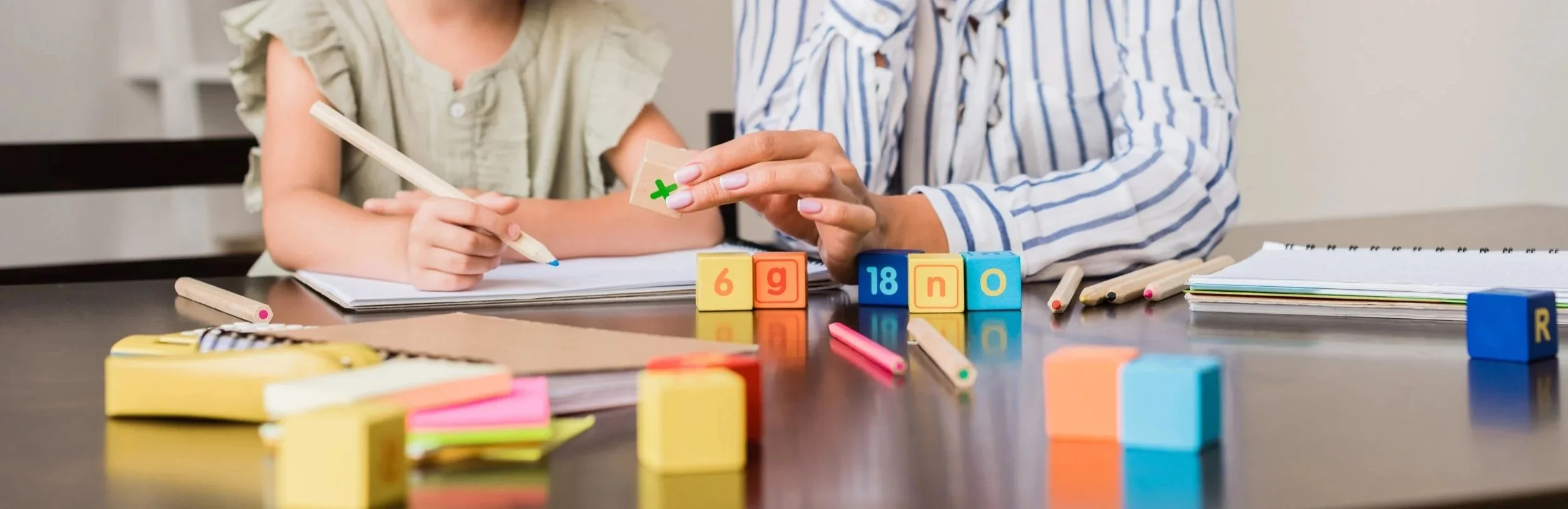 Two children at a table playing with colorful alphabet and number blocks, writing in a notebook, surrounded by pens and paper.