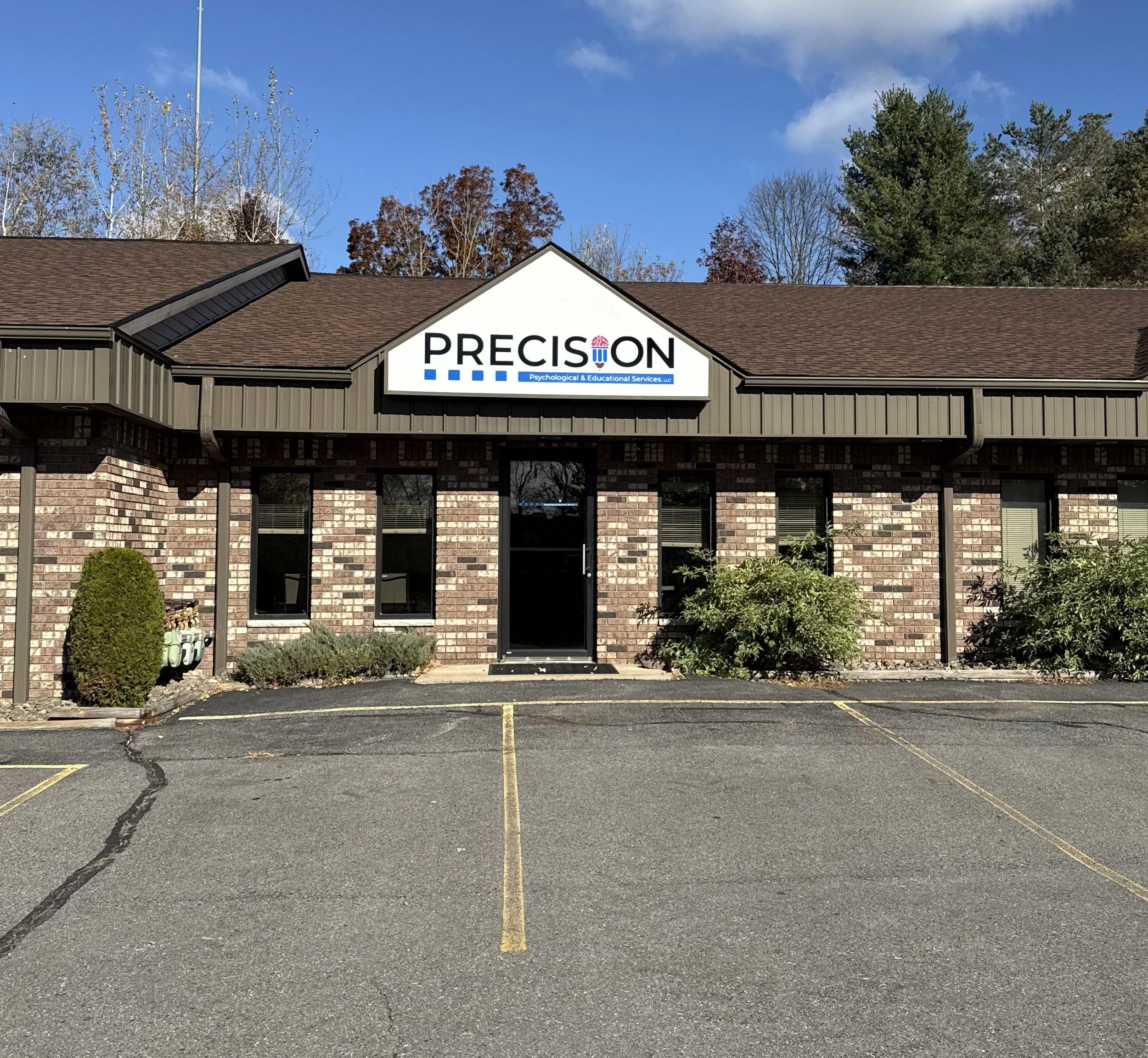 Brick building with a brown roof and a sign that reads 'PRECISION' with additional text about psychological and educational services. There are parking spaces in front and some bushes near the entrance, with a clear blue sky above.