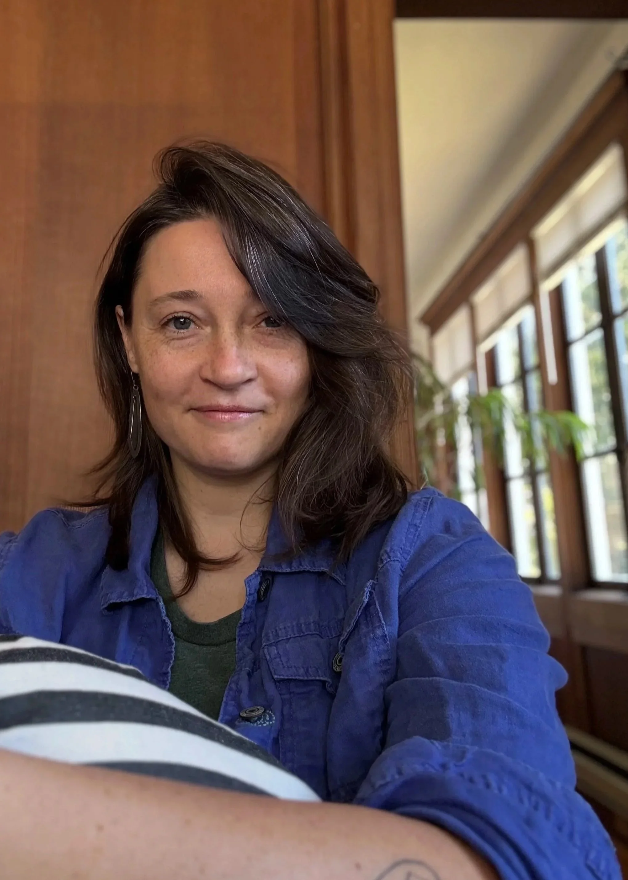 A white woman with freckles and long dark brown hair looks into the camera. She's wearing awesome striped pants and smiling slightly. Behind her is redwood paneling and, in the distance, trees visible through the windows.