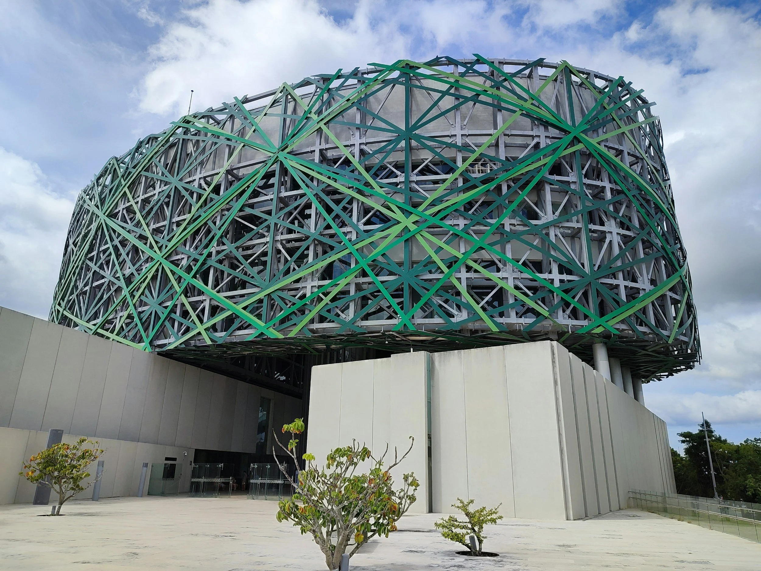 Modern building with a unique, circular, lattice-like green and gray exterior structure, gray concrete base, small trees in foreground, partly cloudy sky in background.