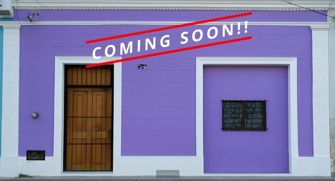 A building painted purple with white trim, a brown wooden door, a window with black bars, and a sign that says "COMING SOON!!" in white letters with red lines above and below.