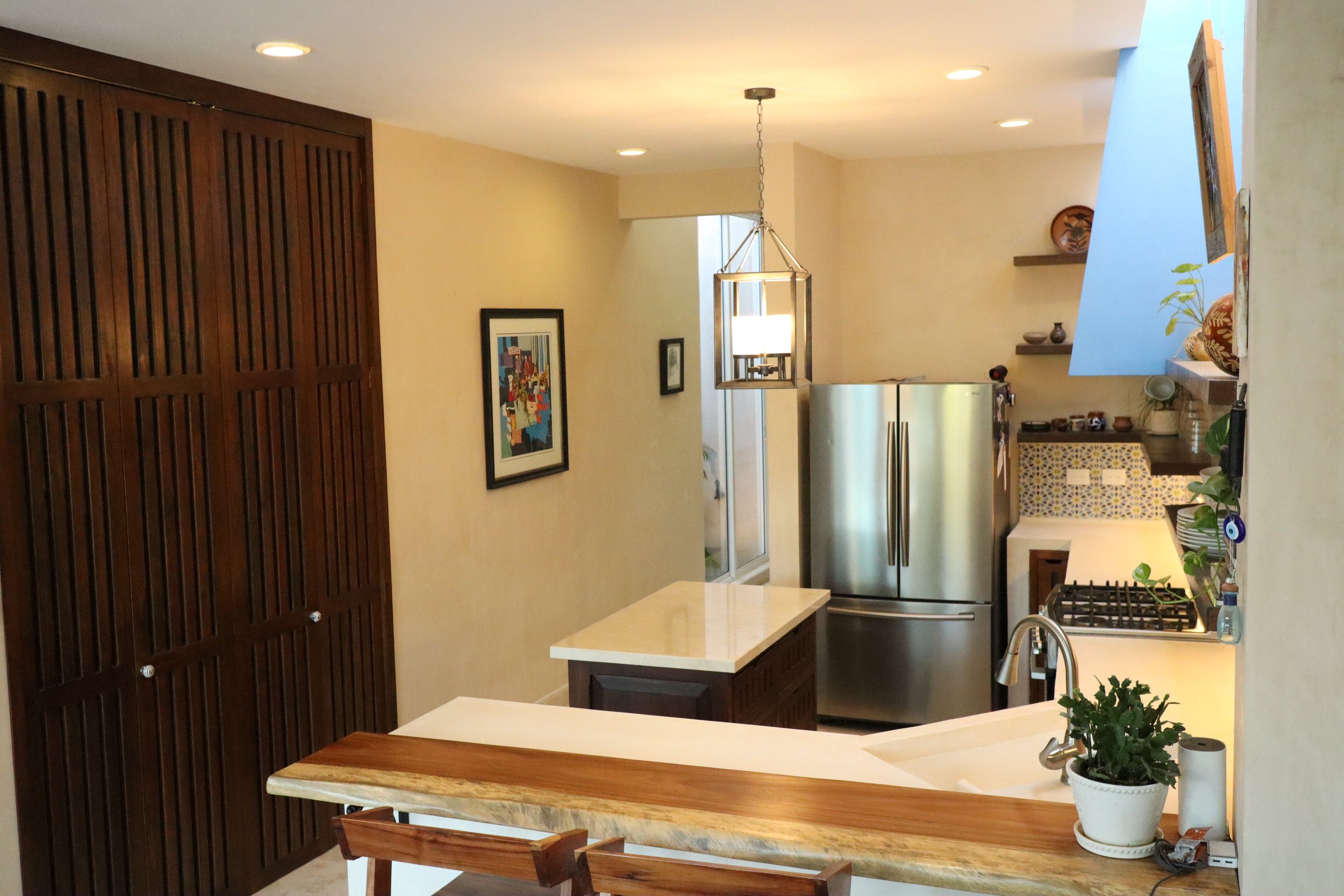 Modern kitchen with beige walls, wooden cabinets, stainless steel refrigerator, white countertops, a small island, and a breakfast bar with a wooden edge. There are hanging lights, shelves with decorative items, and a plant near the sink.