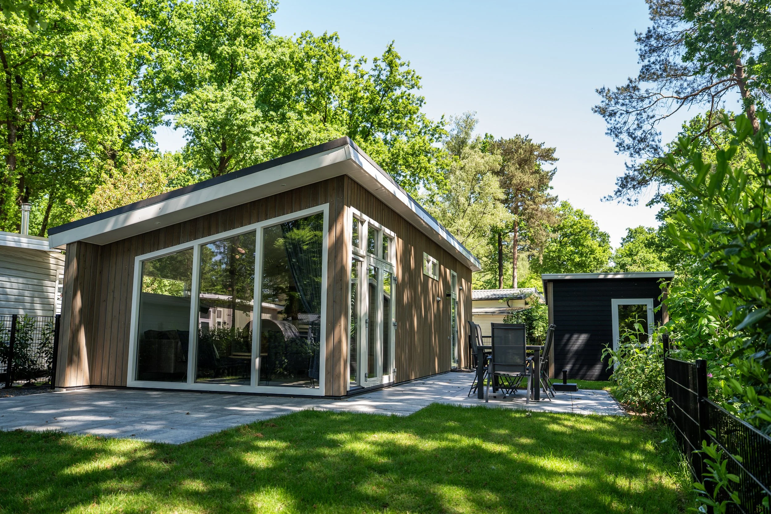 Moderne houten huis met grote glazen ramen en een patio met tuinmeubilair, omringd door groene bomen en een hek.