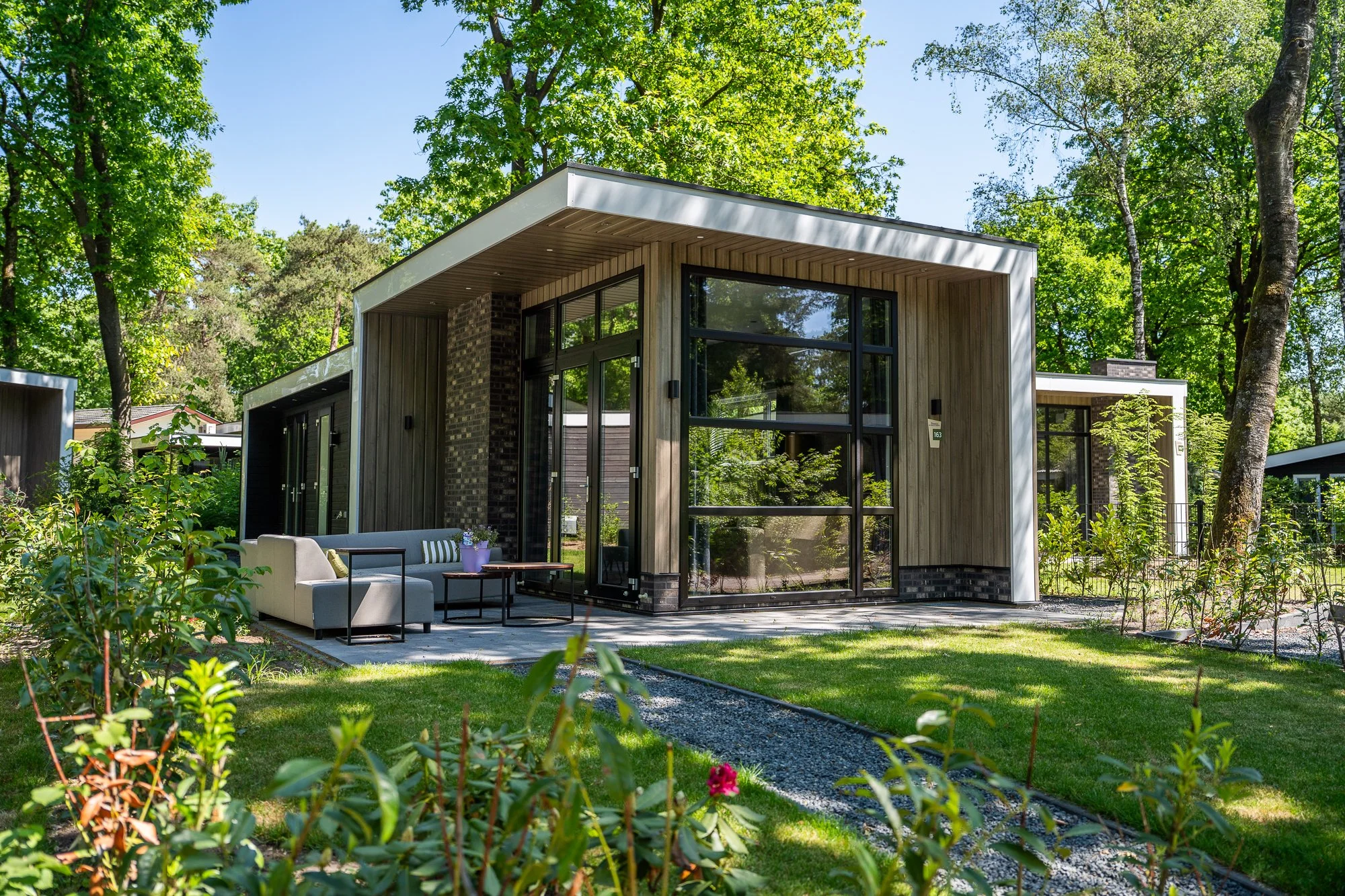 Moderne woning in een groene tuin met zitgedeelte, grote ramen, houten gevel, omringd door bomen en planten.