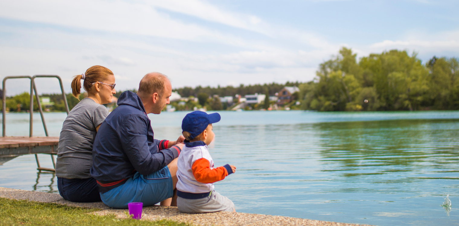 Gezin zit aan de rand van een meer en kijkt naar het water, met een vrolijke sfeer en groene bomen op de achtergrond.