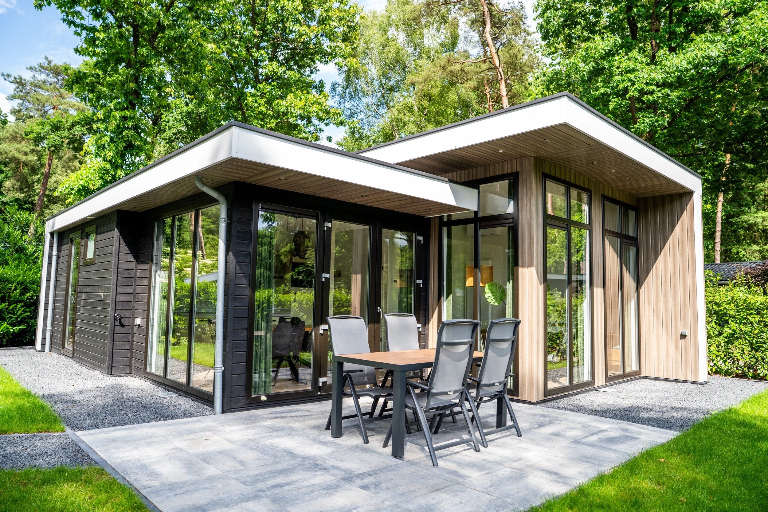 Moderne woning met grote glazen ramen en een buitentafel met zes stoelen op een terras, omgeven door grote groene bomen.