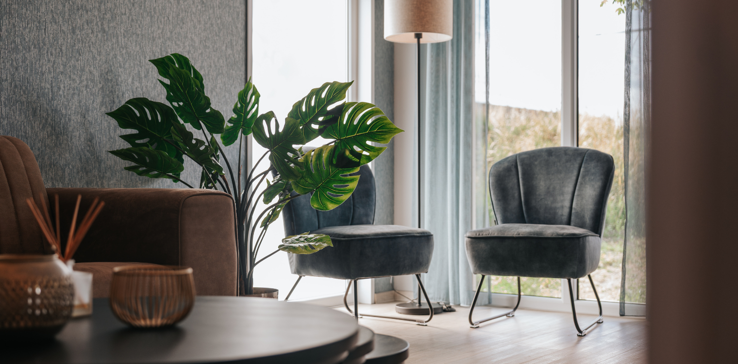 Modern woonkamer met grote plant, twee grijze stoelen met spatels, vloer met houten vloer, grote ramen met uitzicht op de natuur, schemerige verlichting.