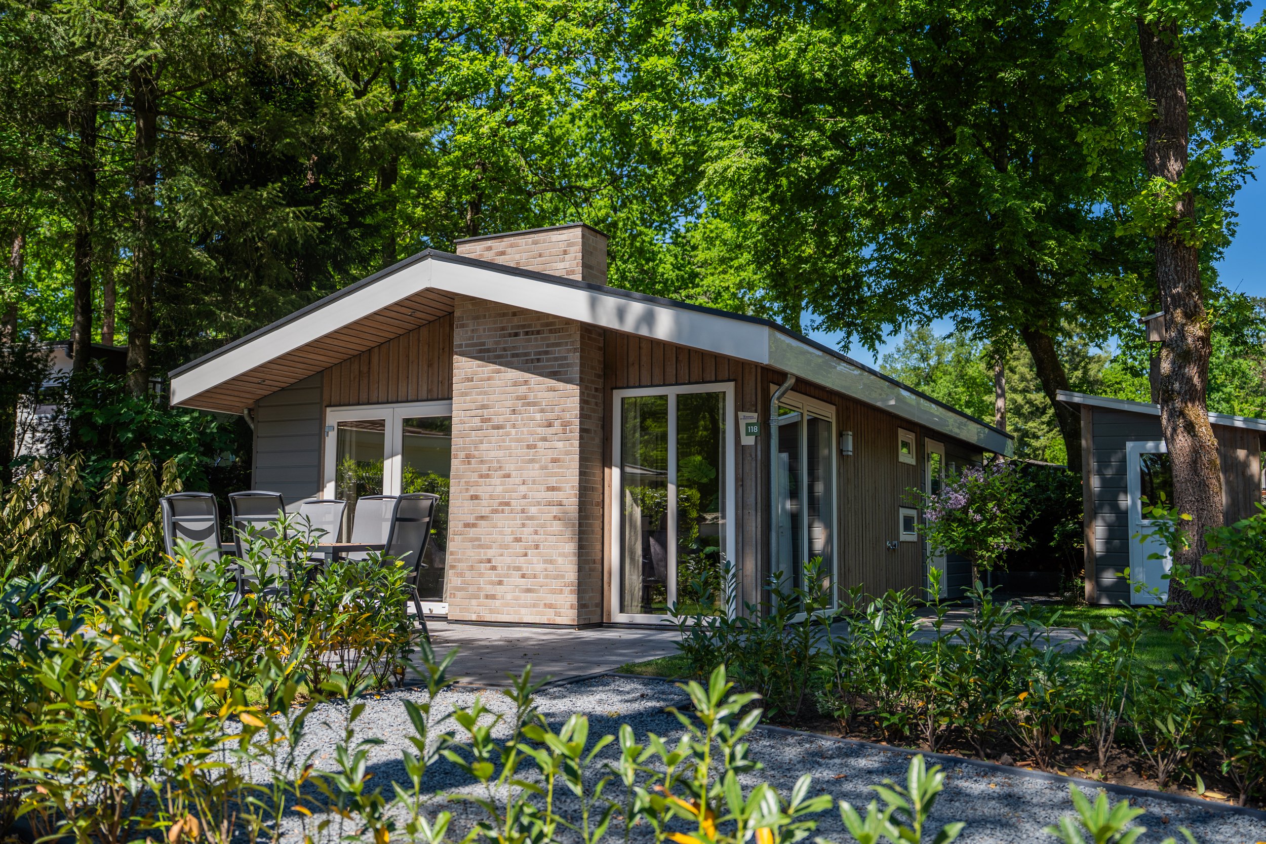 Een modern eenlaagshuis met een houten gevel en grote glazen deur, omgeven door groene bomen en struiken, met een zithoek op een terras.