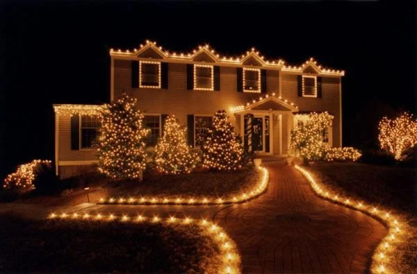 A house decorated with Christmas lights, including lights outlining the roof, windows, and pathway, with decorated trees in the yard at night for Christmas lighting inspiration for Neon Noel customers in Paris, Texas