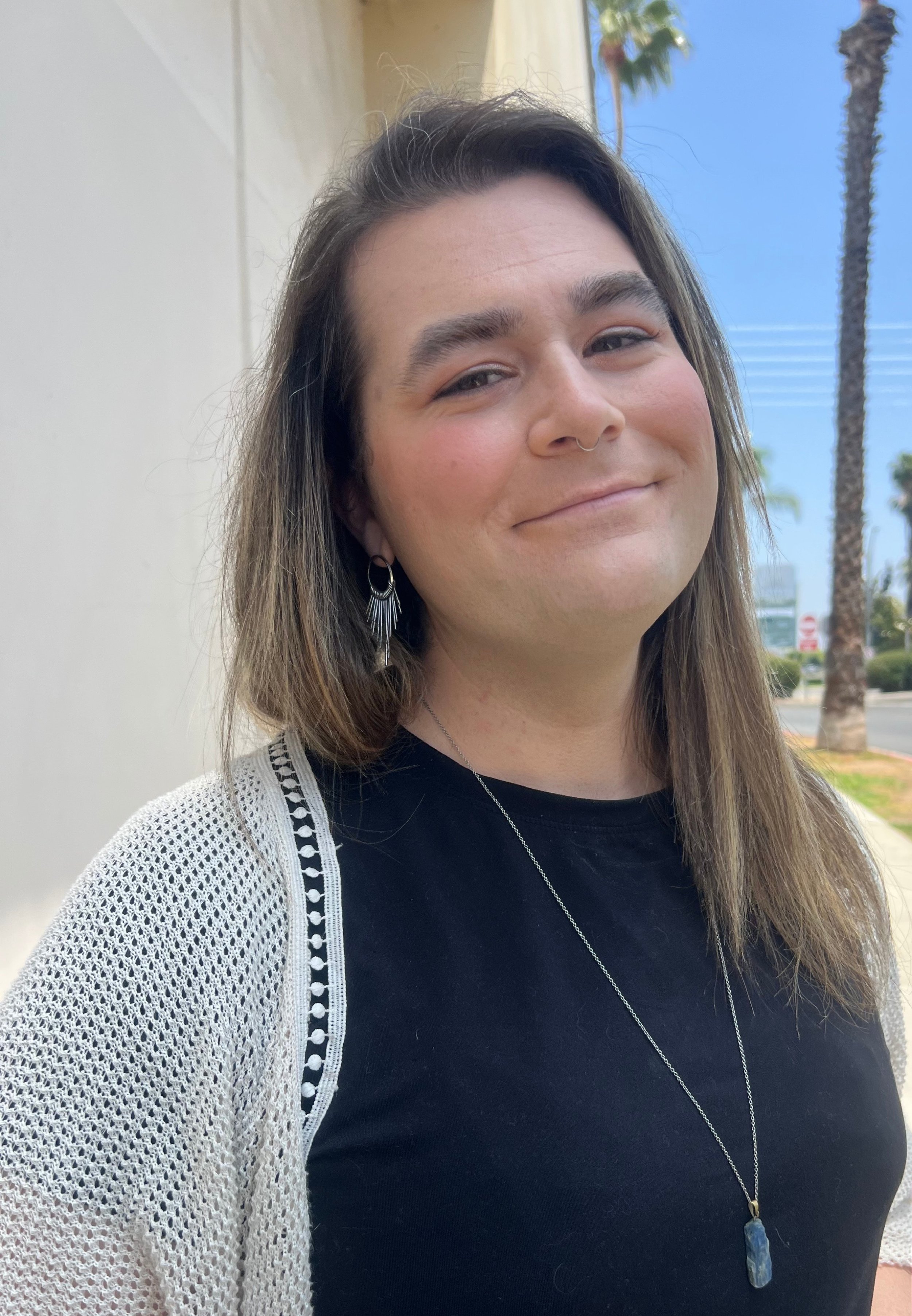 A woman with shoulder-length brown hair, wearing earrings, a septum ring, a black shirt, a white knit cardigan, and a necklace with a blue stone, standing outdoors with palm trees and a street in the background.