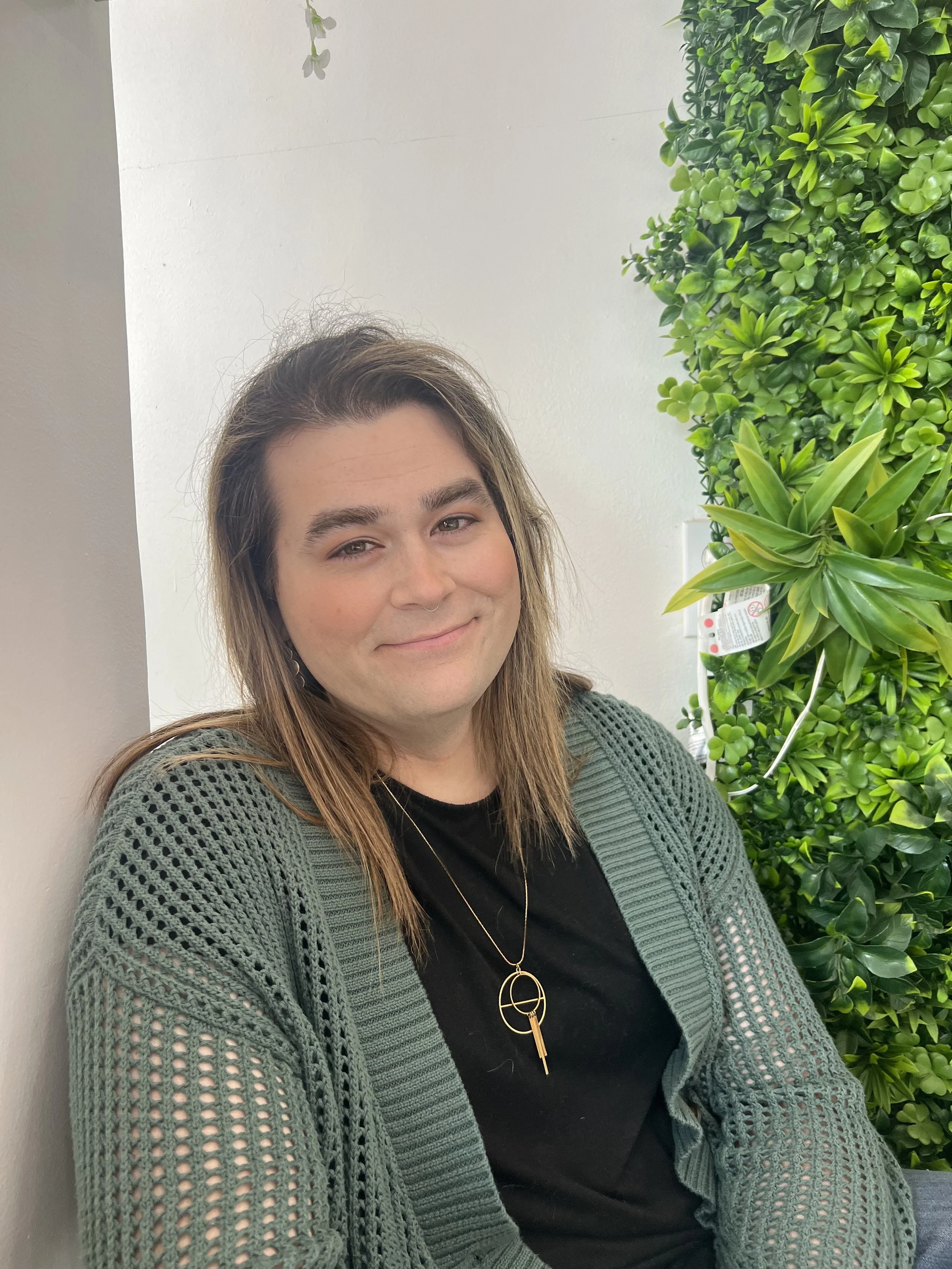 A woman with shoulder-length brown hair and a septum piercing, wearing a black shirt, a green knitted cardigan, and a gold necklace with a circular pendant and a bar, is sitting and smiling at the camera. She is seated next to a white wall and a lush green wall of plants.