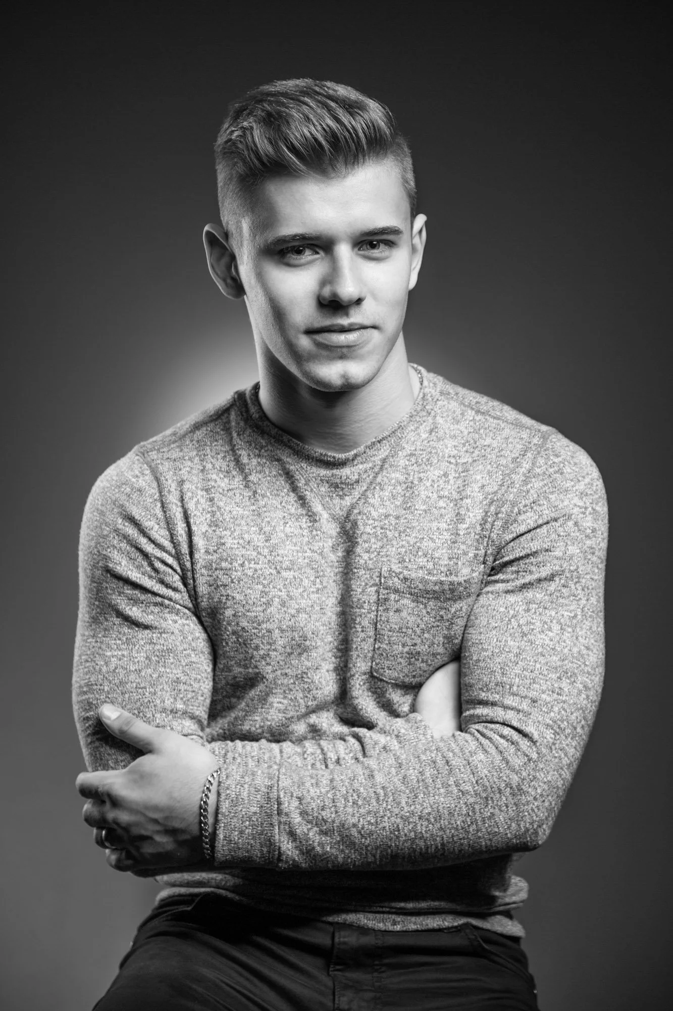 Portrait, Fitness & Commercial Photographer Zurich. Black and white portrait of a young man with short, styled hair, wearing a long-sleeved knit sweater and a chain bracelet, sitting against a plain background.