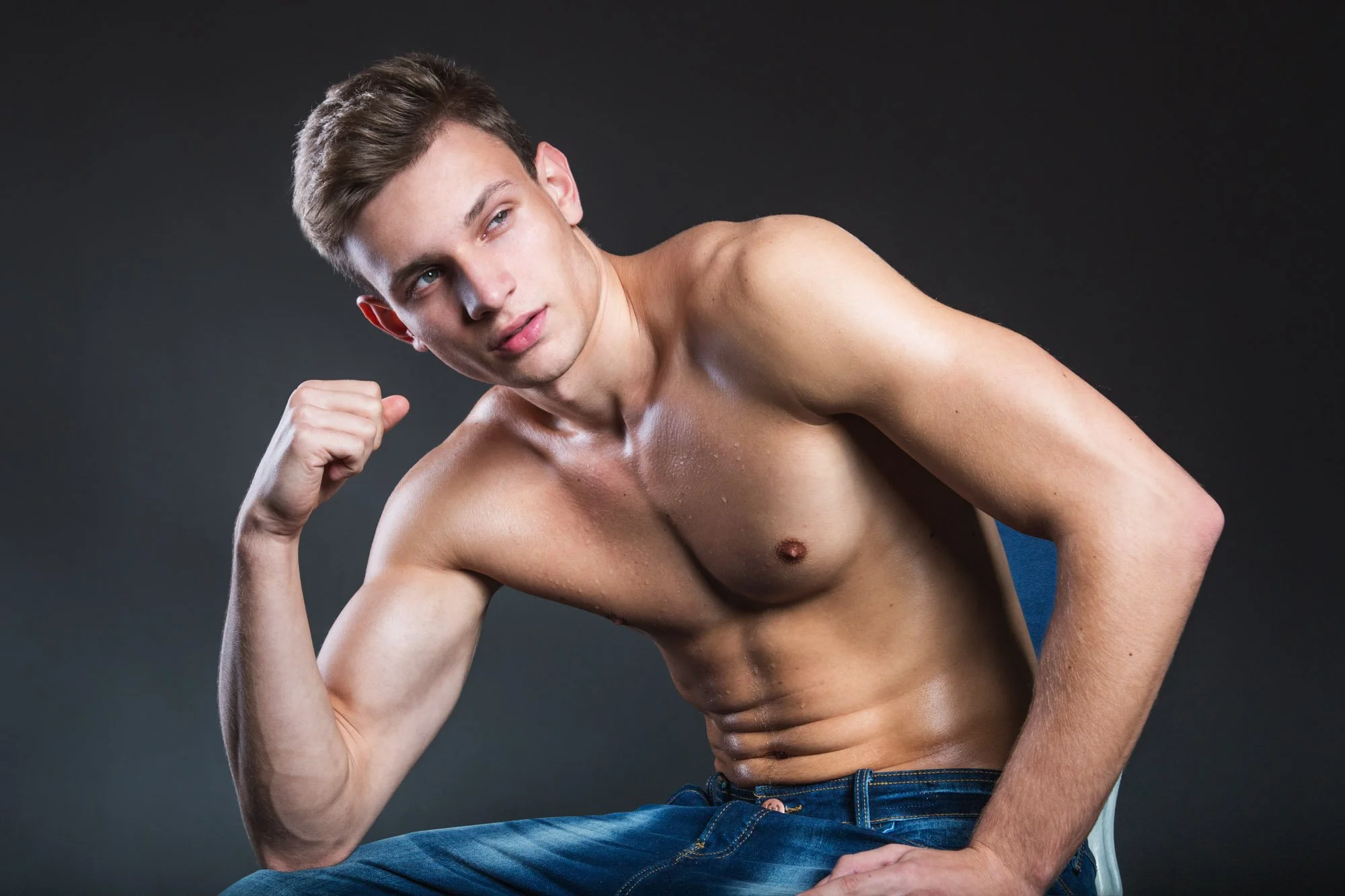 A young man with short brown hair and blue eyes is shirtless, showing his toned upper body. He is sitting on a white chair against a plain dark background, with his right arm raised and fist clenched, striking a confident pose.