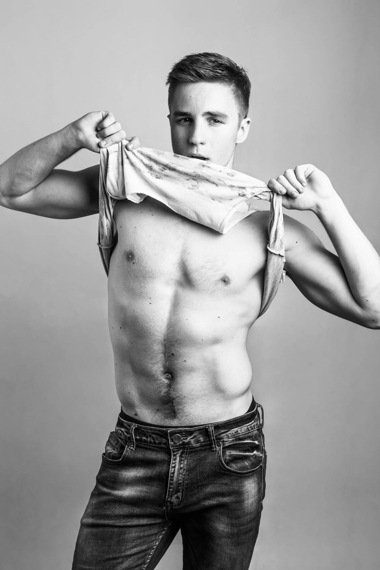 Black and white photo of a shirtless young man with short hair, holding a wrinkled t-shirt in front of his face, standing against a plain background.