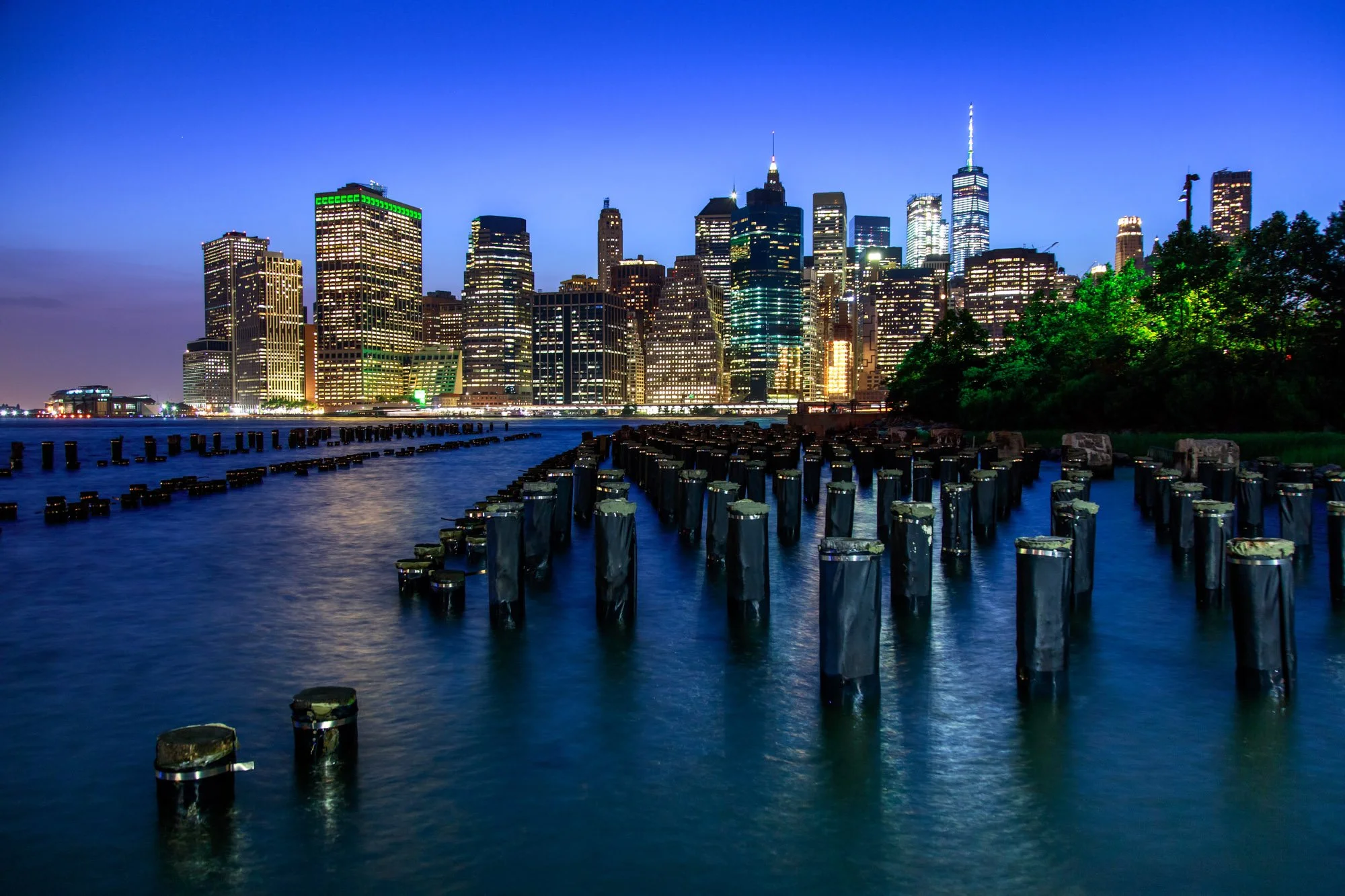 Nighttime view of New York City skyline with illuminated skyscrapers, reflections on the water, and old wooden pilings in the foreground.