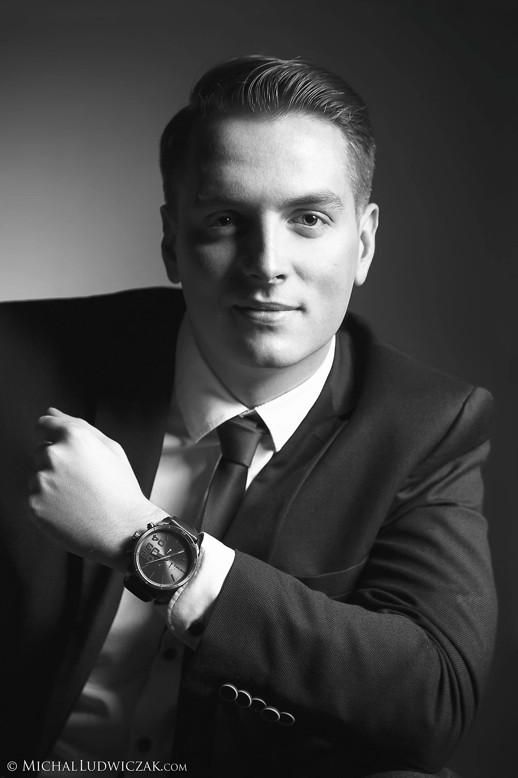Black and white portrait of a young man wearing a suit, tie, and watch, smiling subtly, with one arm resting on his lap.