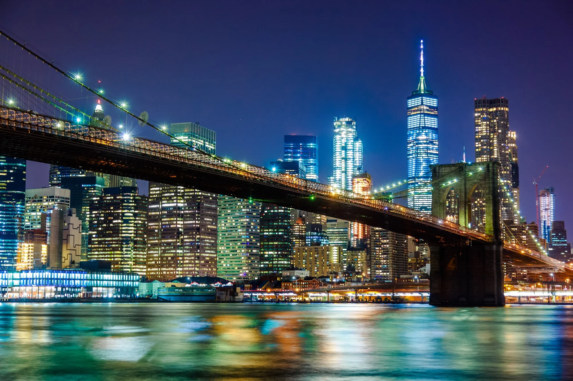 Fine art print photography for modern living roNighttime view of New York City skyline featuring the Empire State Building illuminated in blue and white, with other skyscrapers lit up and reflecting on the water below, and a bridge in the foreground.