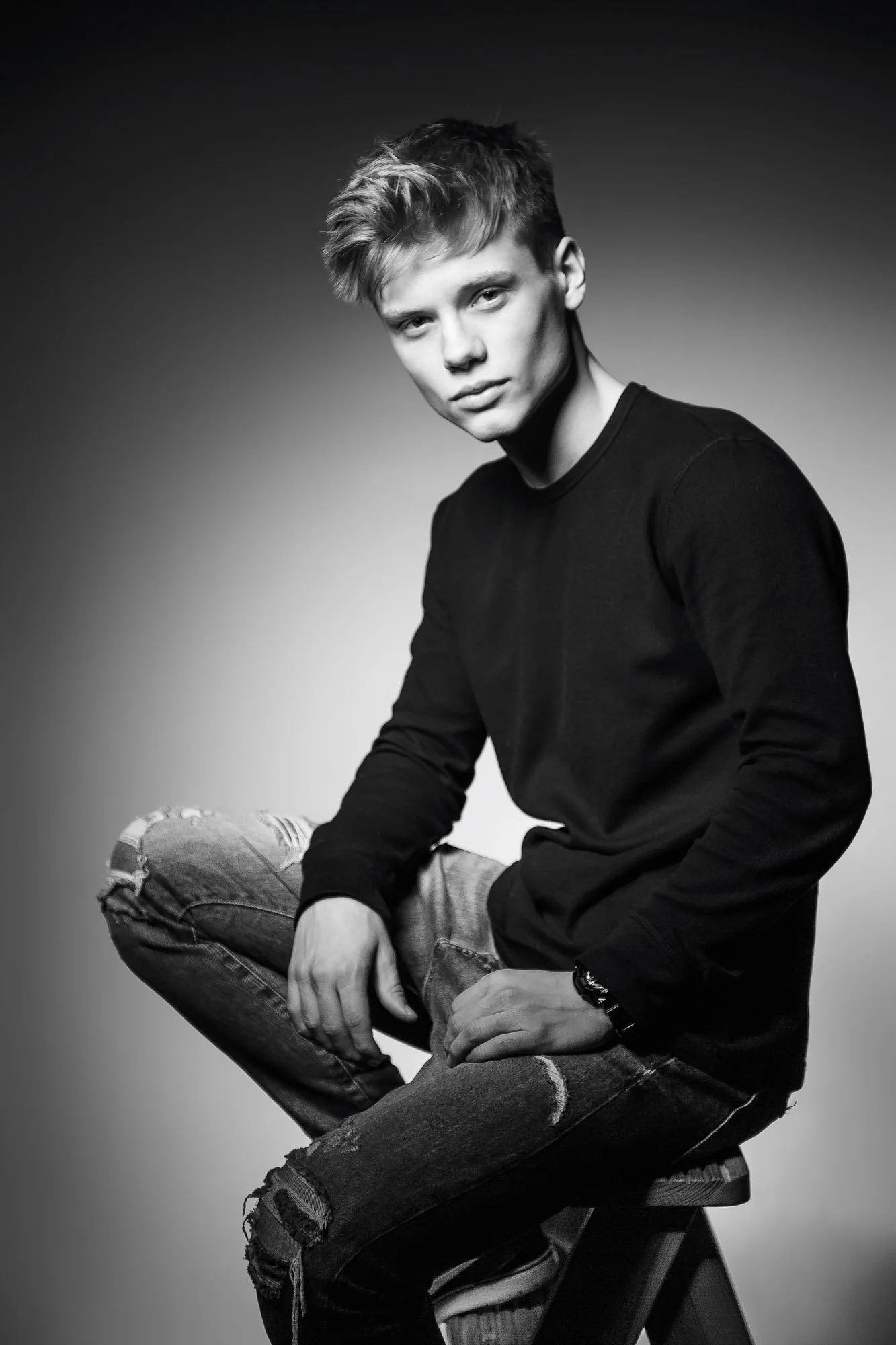 A black and white portrait photograph of a young man with short messy hair, sitting on a stool. He is wearing a black long-sleeve shirt and ripped jeans, and has a serious expression with a slight squint. The background is plain and gradient, fading 