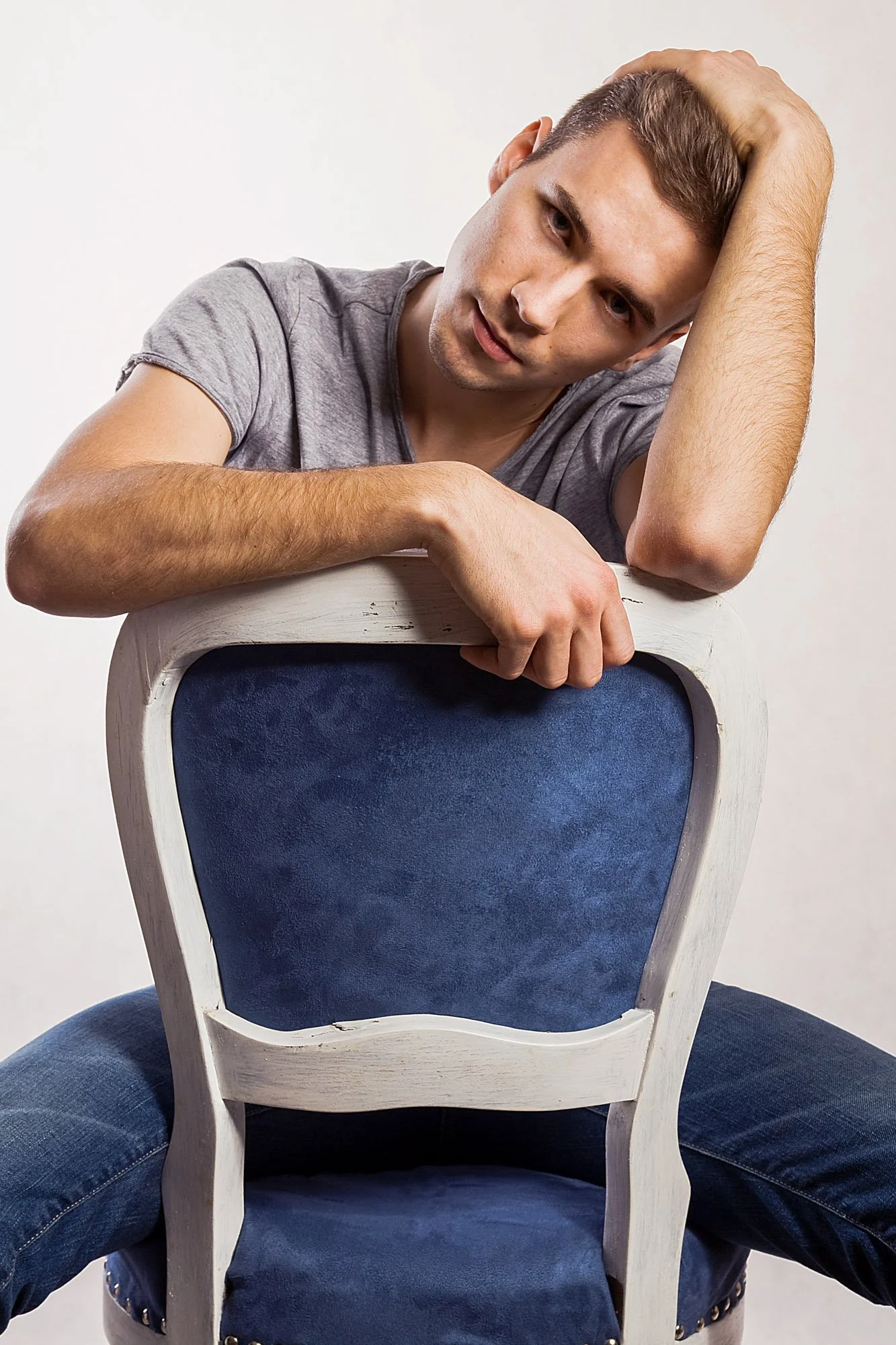 A young man in a gray t-shirt and jeans sitting backwards on a white wooden chair with a blue cushion. He has brown hair and is leaning his head on his left hand, looking at the camera with a thoughtful expression. The background is plain white.