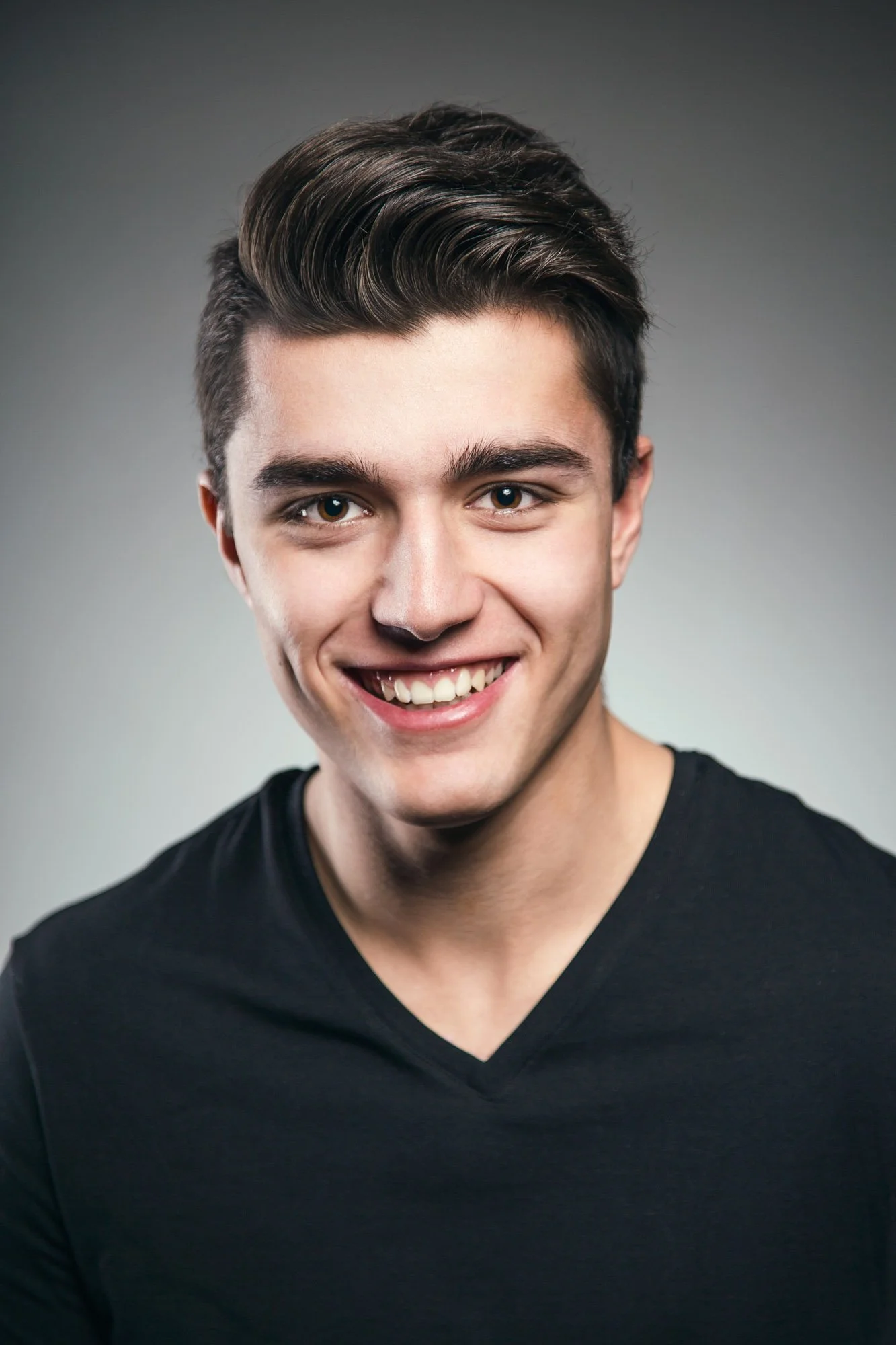 Artistic male portrait photography Zurich. Close-up portrait of a young man with dark hair styled to the side, smiling and wearing a black V-neck shirt against a gray background.