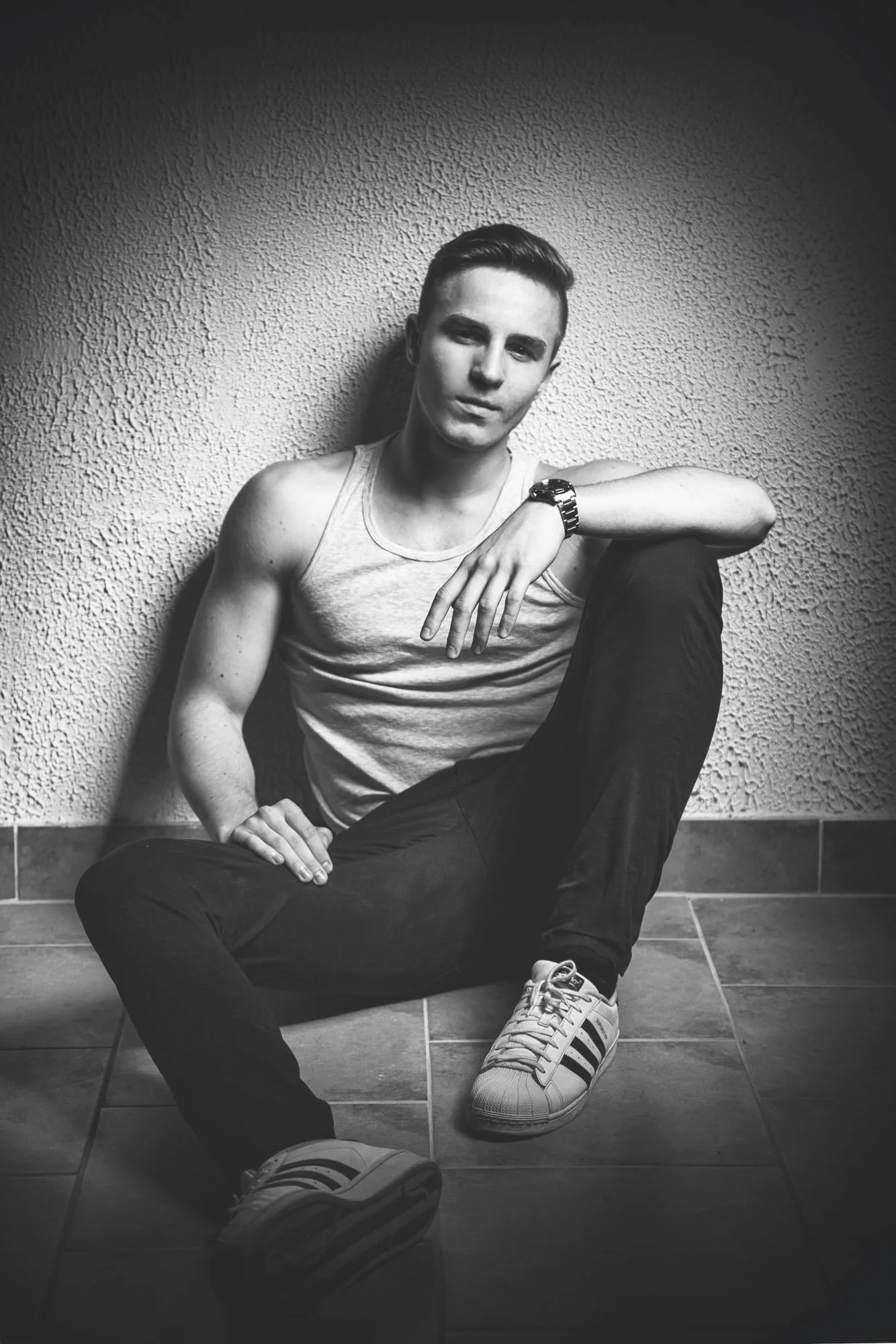 Portrait, Fitness & Commercial Photographer ZurichBlack-and-white photo of a young man sitting on the floor against a textured wall, with one arm resting on his knee and wearing casual clothing, including a sleeveless shirt, dark pants, and sneakers.