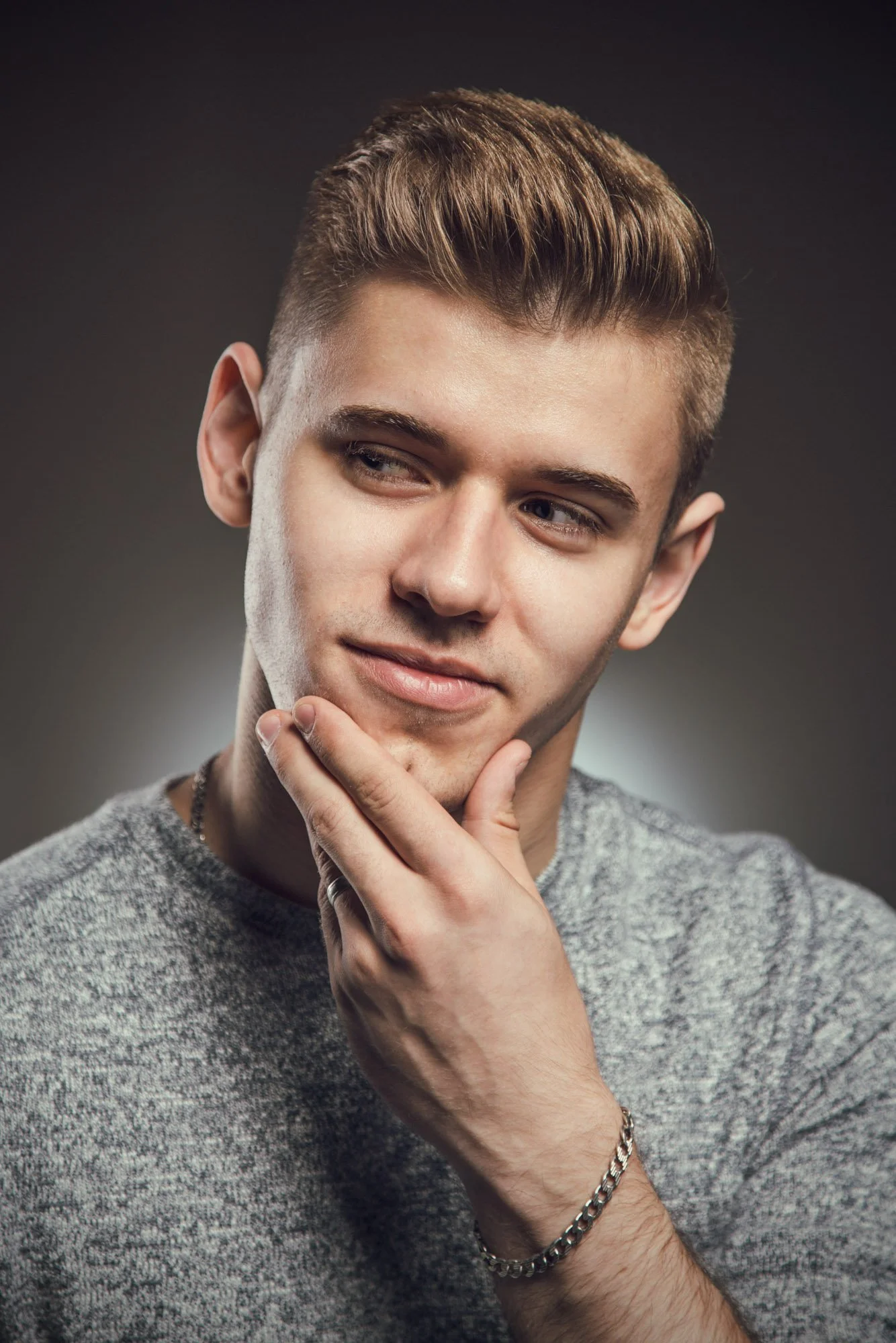 Portrait, Fitness & Commercial Photographer Zurich. A young man with styled light brown hair posing against a gray background, wearing a gray sweater, a chain bracelet, and a ring, with his hand touching his chin and looking to the side.