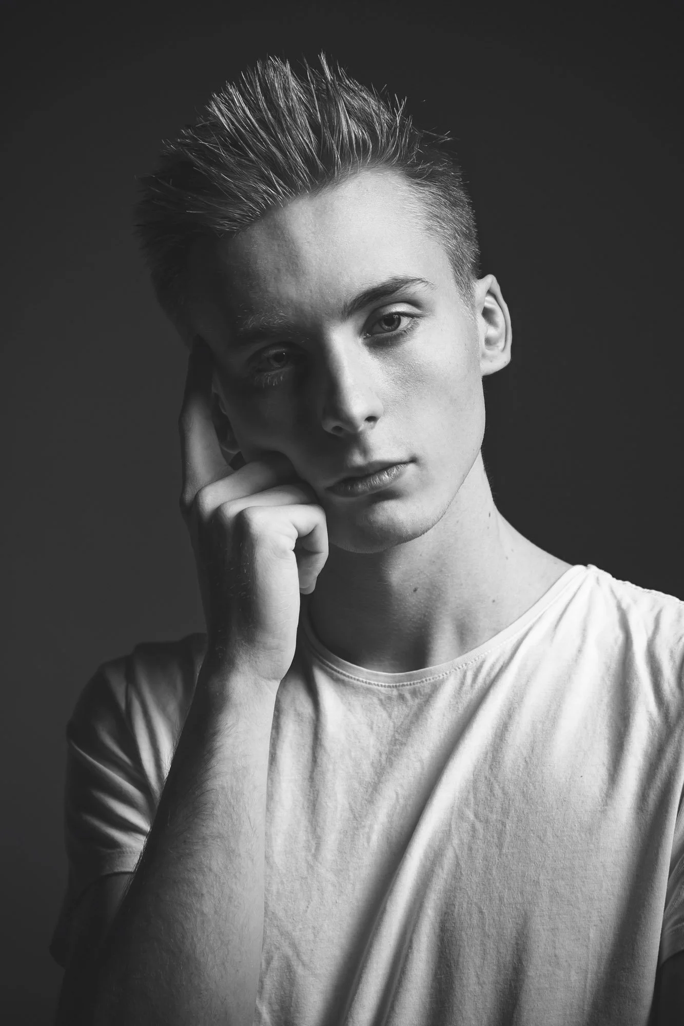 Black and white portrait of a young person with short, styled hair, wearing a light-colored shirt, resting their face on one hand, looking directly at the camera with a neutral expression.