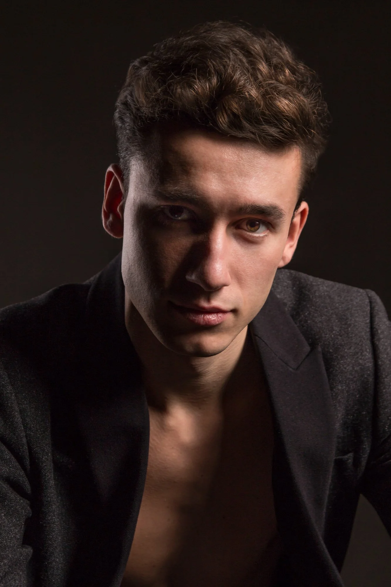 A young man wearing a dark blazer with an open shirt reveals part of his chest, set against a dark background.