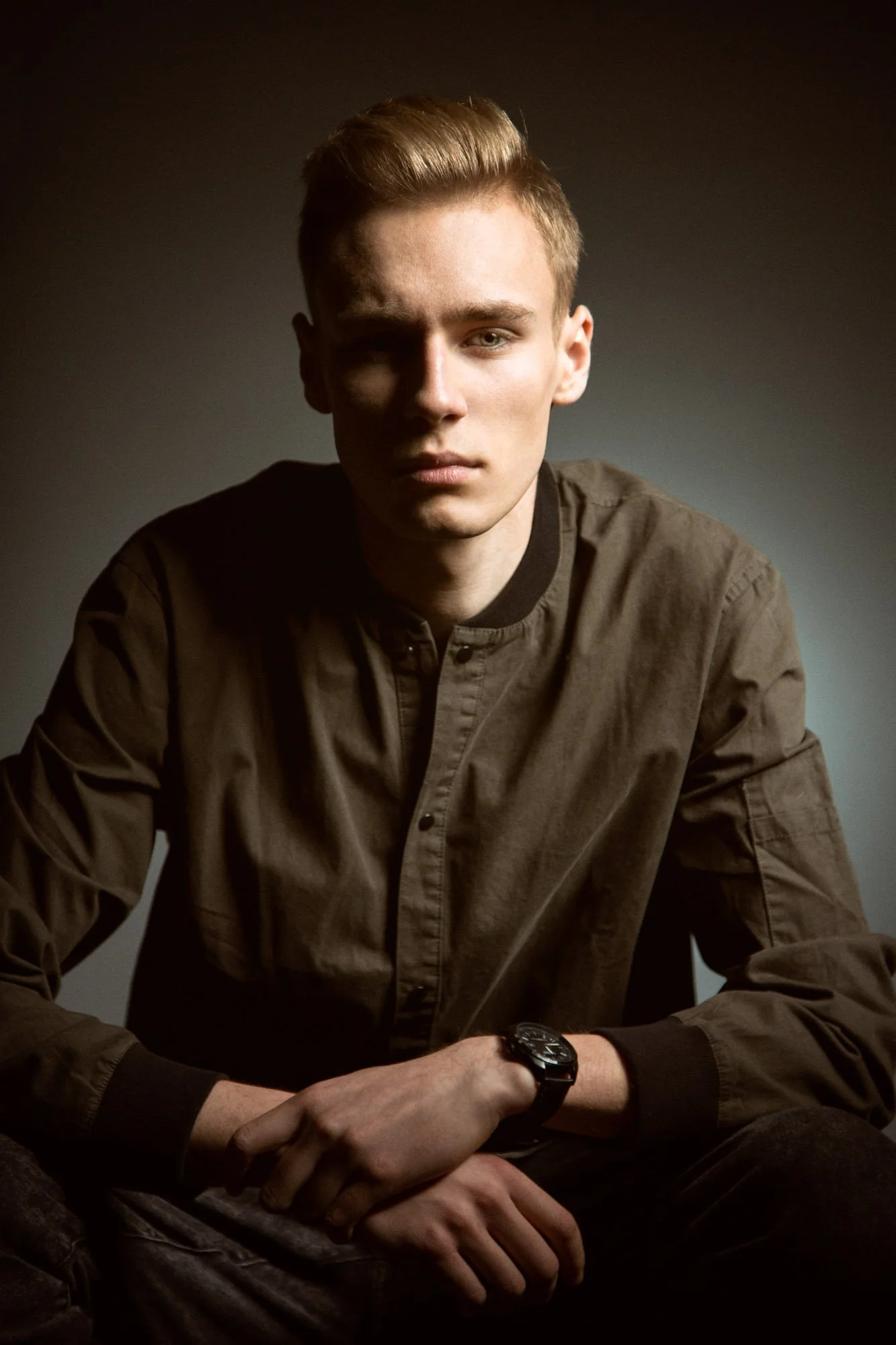 Young man with blonde hair and blue eyes sitting against a dark background, wearing a brown shirt and black watch.
