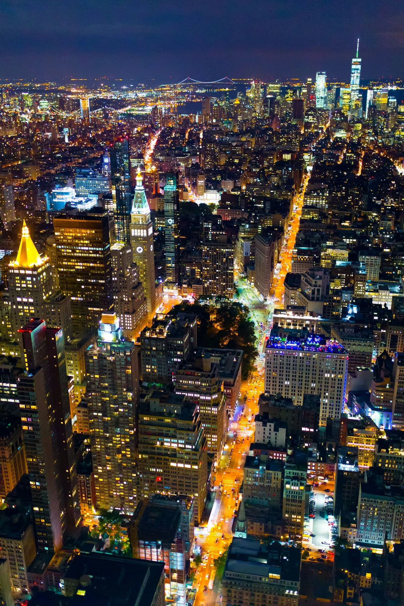 Aerial night view of a city skyline with illuminated skyscrapers, streets, and bridges.