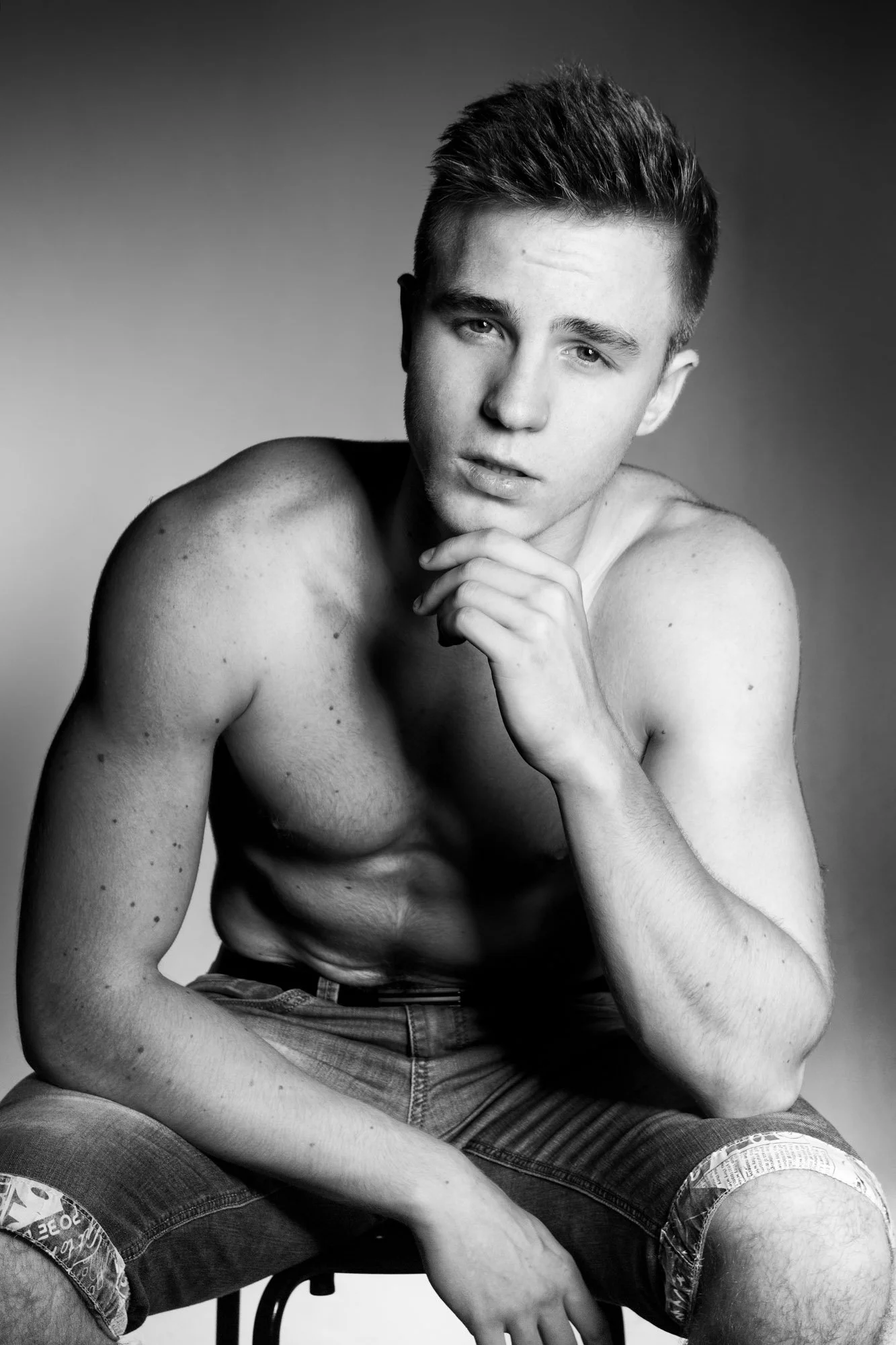 Black and white photo of a young man with short, styled hair and no shirt, sitting with one hand resting on his chin. He is wearing denim shorts and mesh shorts underneath, with a serious expression and a subtle gaze.